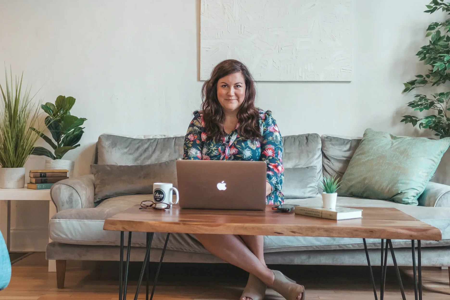 A woman sitting on a sofa in a cozy living room, working on a laptop placed on a wooden coffee table with books and potted plants.