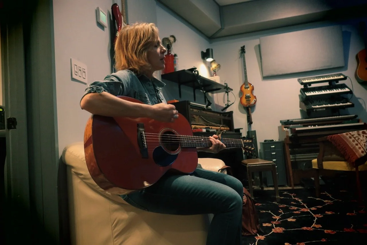 Woman playing acoustic guitar in a music studio with various instruments and equipment.