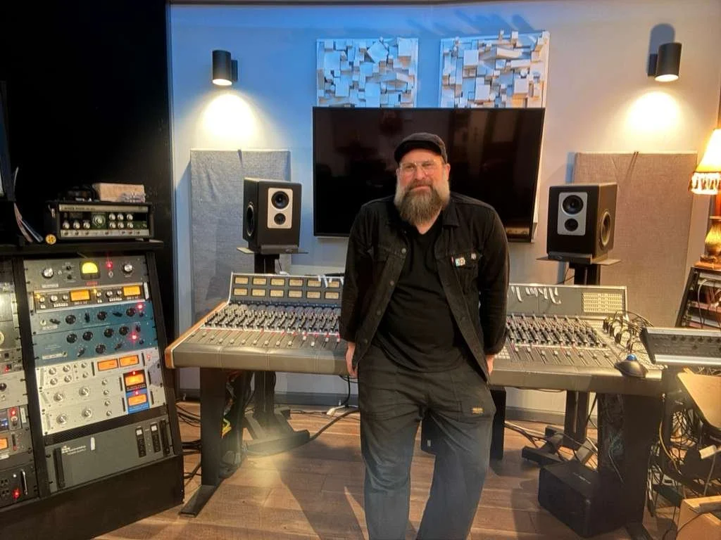 A man with a beard and glasses standing in a recording studio, surrounded by audio equipment including a mixing console, studio monitors, and racks of audio gear.