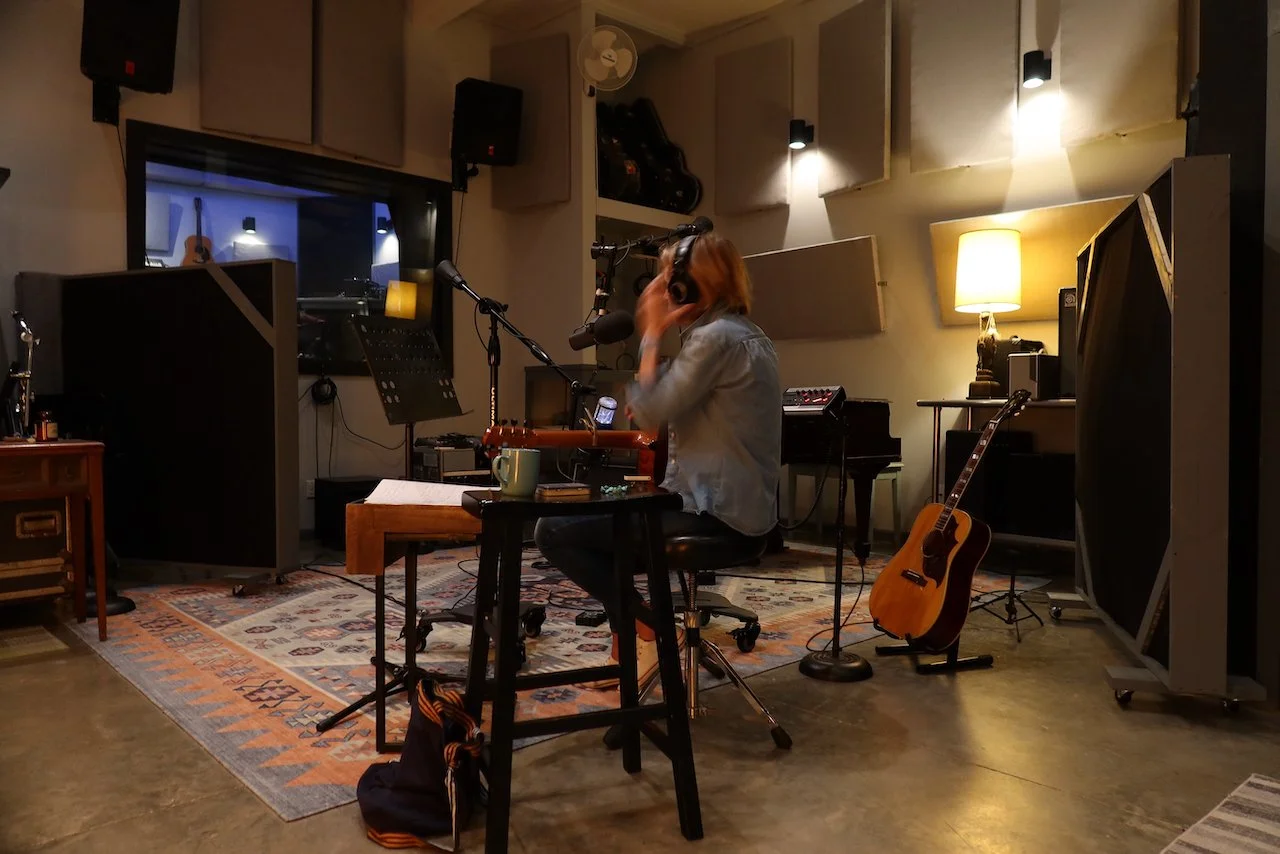Woman in a recording studio, sitting at a desk with a guitar, wearing headphones, with microphones and music equipment around, illuminated by warm lighting.