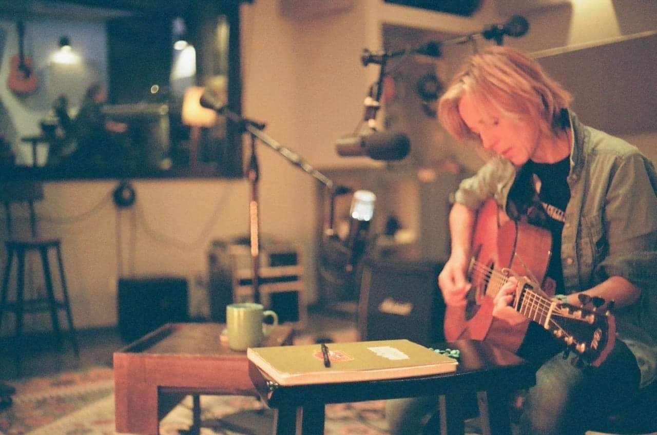 A woman playing an acoustic guitar in a cozy room with recording equipment and guitars on the wall.
