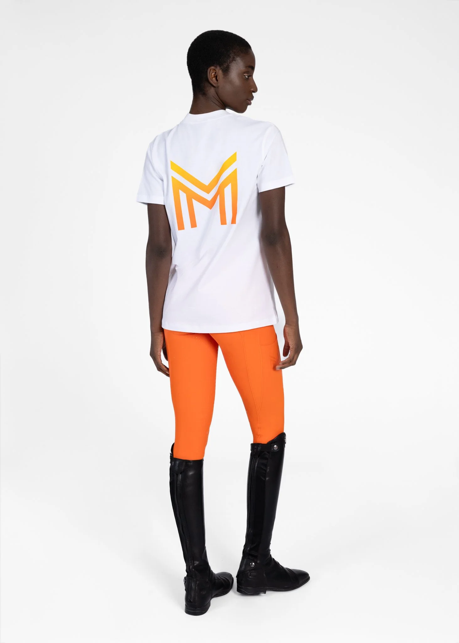 A person with dark skin and short hair wearing a white T-shirt with an orange geometric logo on the back, orange leggings, and black knee-high boots, standing against a plain white background.