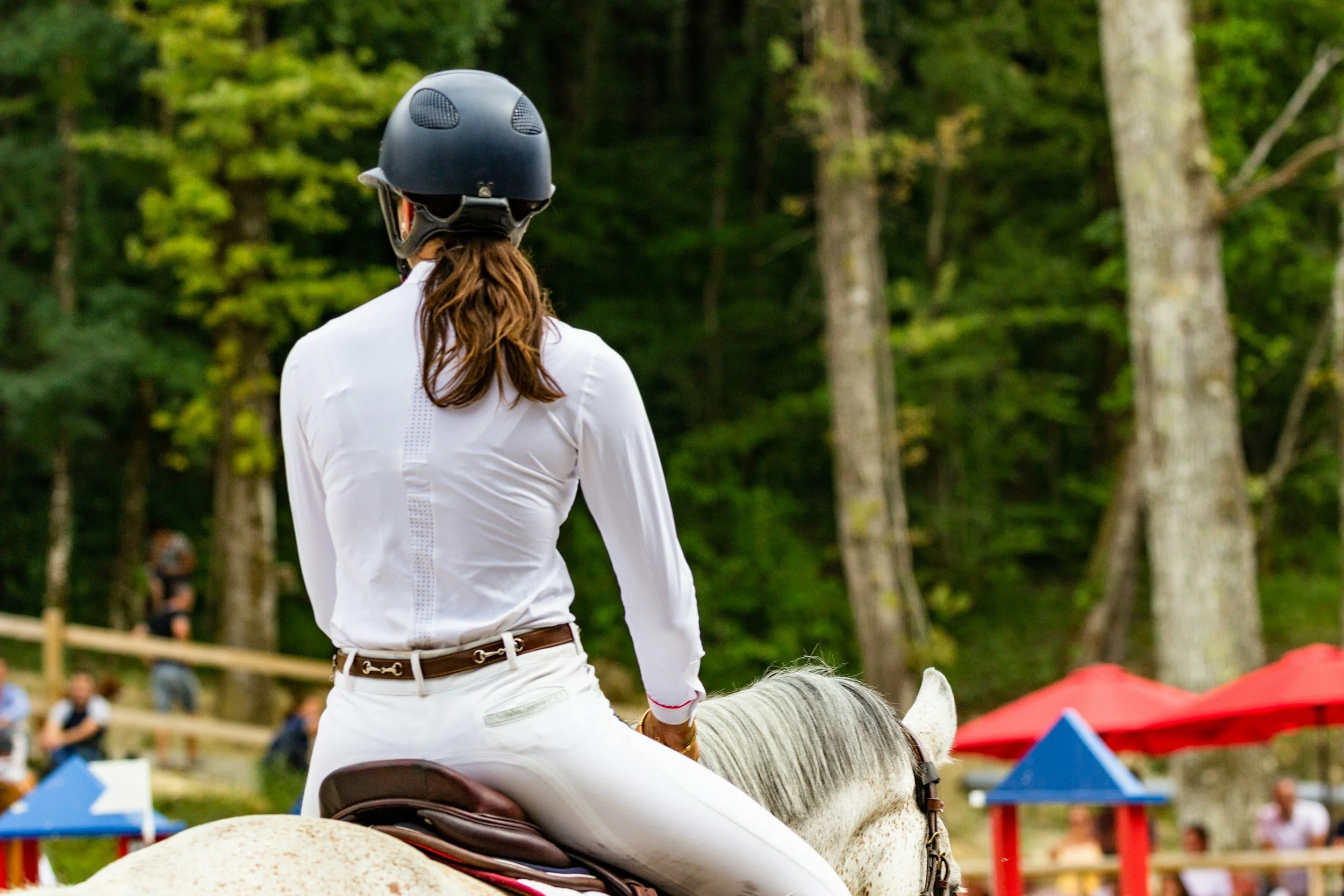 Woman riding a white horse through a lush green outdoor setting with trees and spectators.