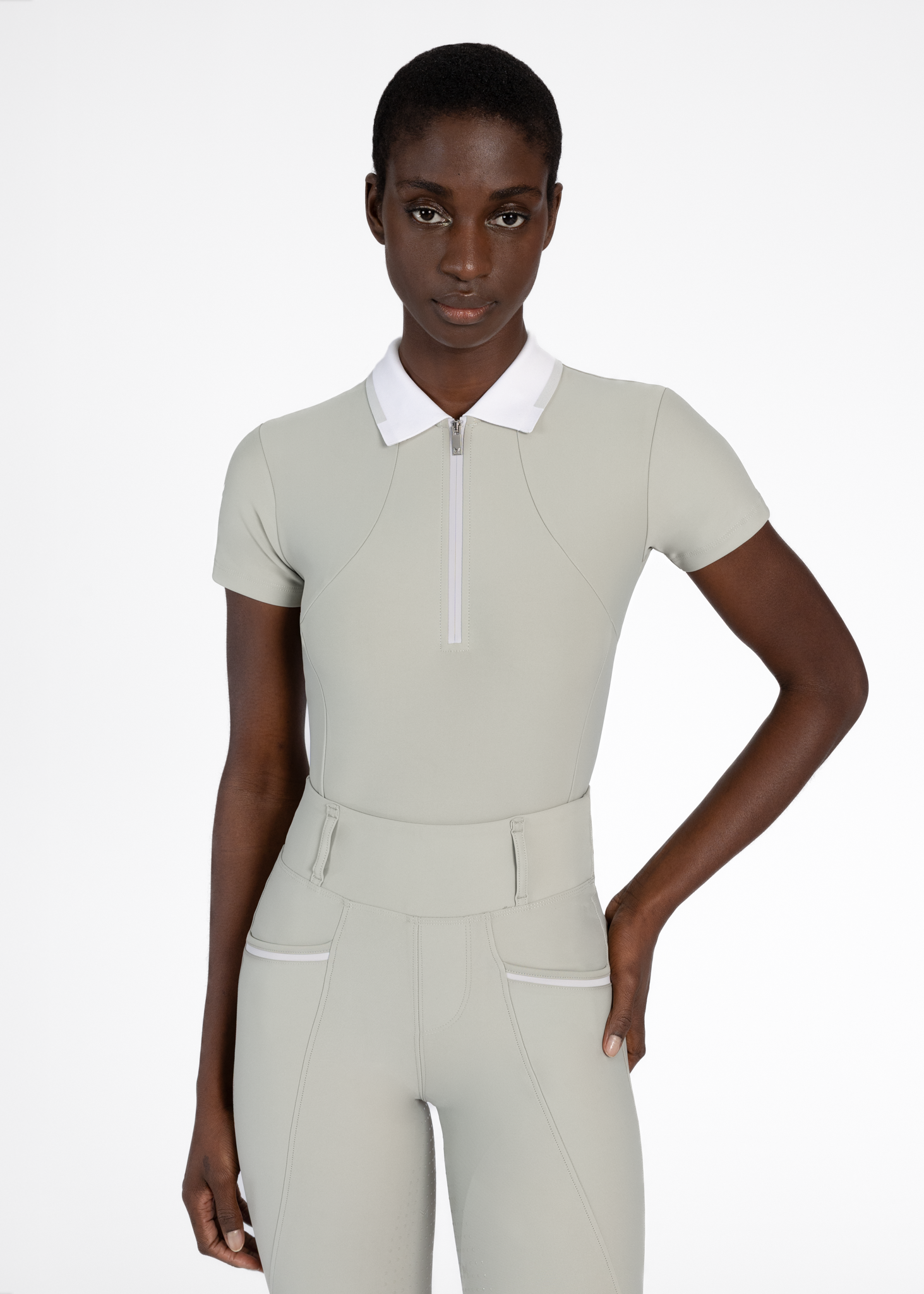 A woman with dark skin and short black hair wearing a light gray, athletic top with a white collar and a zippered front, and matching light gray pants, standing against a plain white background.