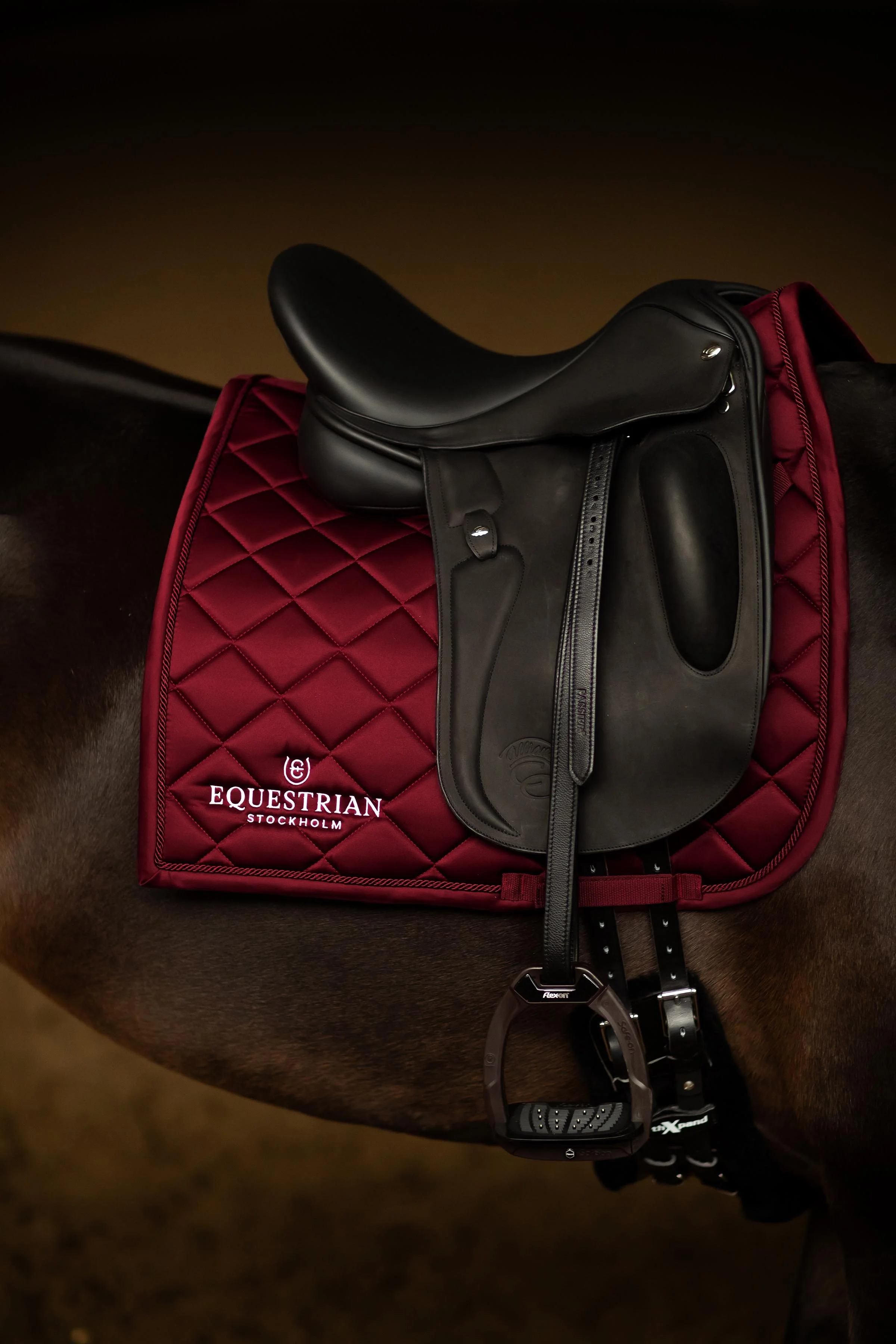 Close-up of a black horse saddle placed on a red quilted saddle pad with 'Equestrian Stockholm' embroidered on it. The saddle pad is on the horse's back, with a black girth underneath. There are also black riding stirrup leathers and a stirrup, along with a black riding helmet with a brand logo hanging from the saddle.