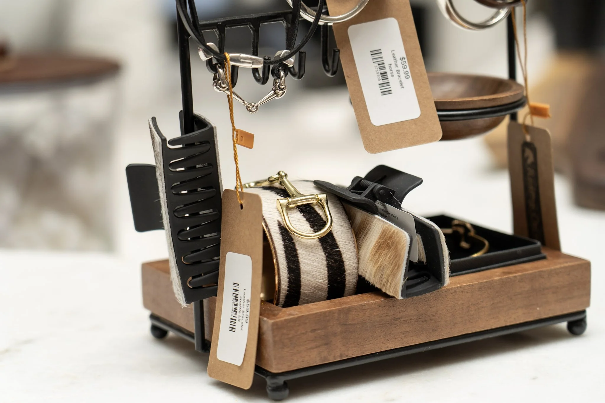 A display of jewelry and accessories including a gold buckle, a black comb, and a footwear item with black and beige stripes, all hanging on a black stand with price tags, set on a wooden base.