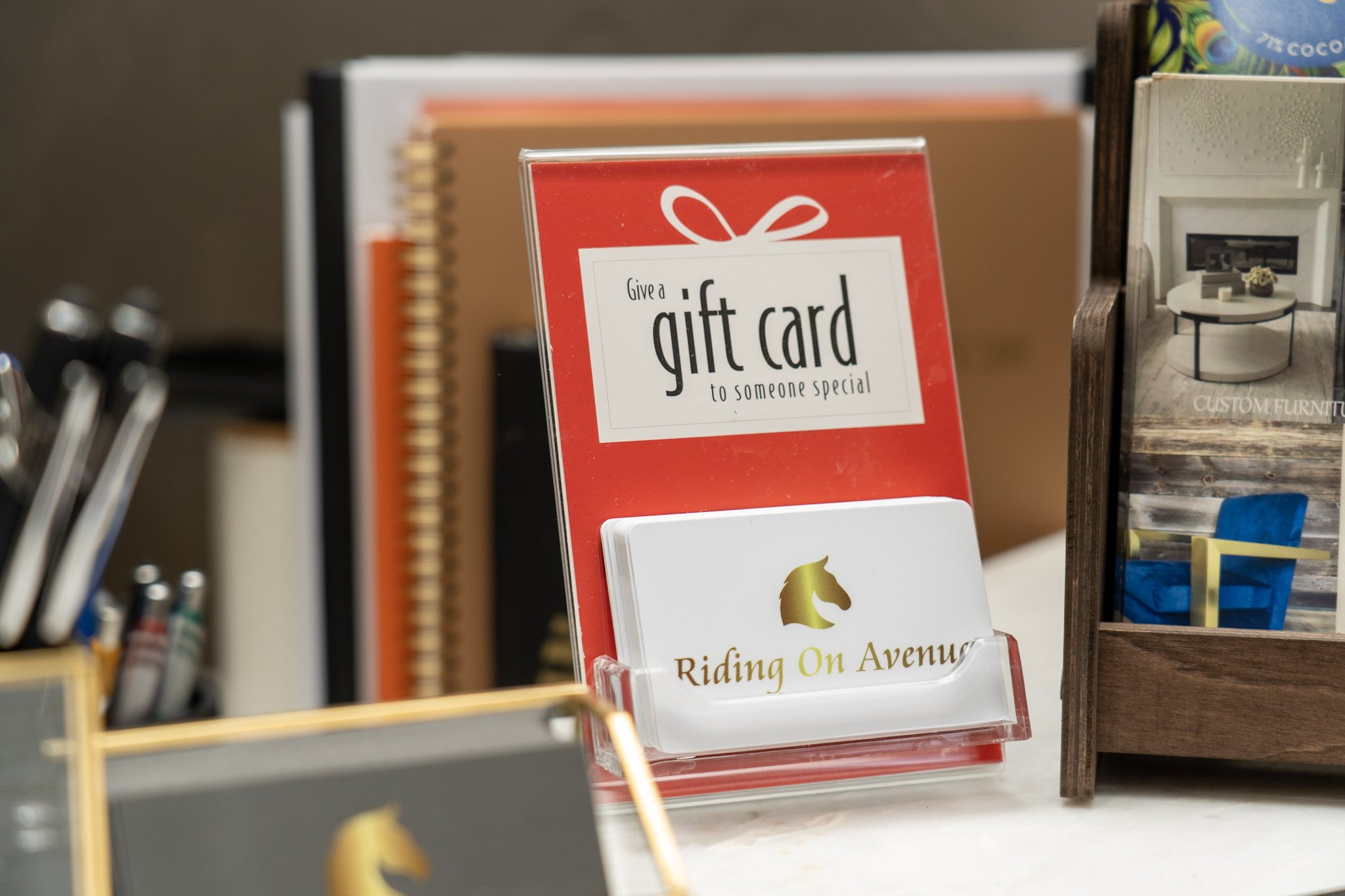 A red and white sign promoting gift cards with the message 'Give a gift card to someone special.' Behind the sign, there are shelves with various books and items, including a small framed picture with a horse silhouette and the words 'Riding On Avenue' underneath.