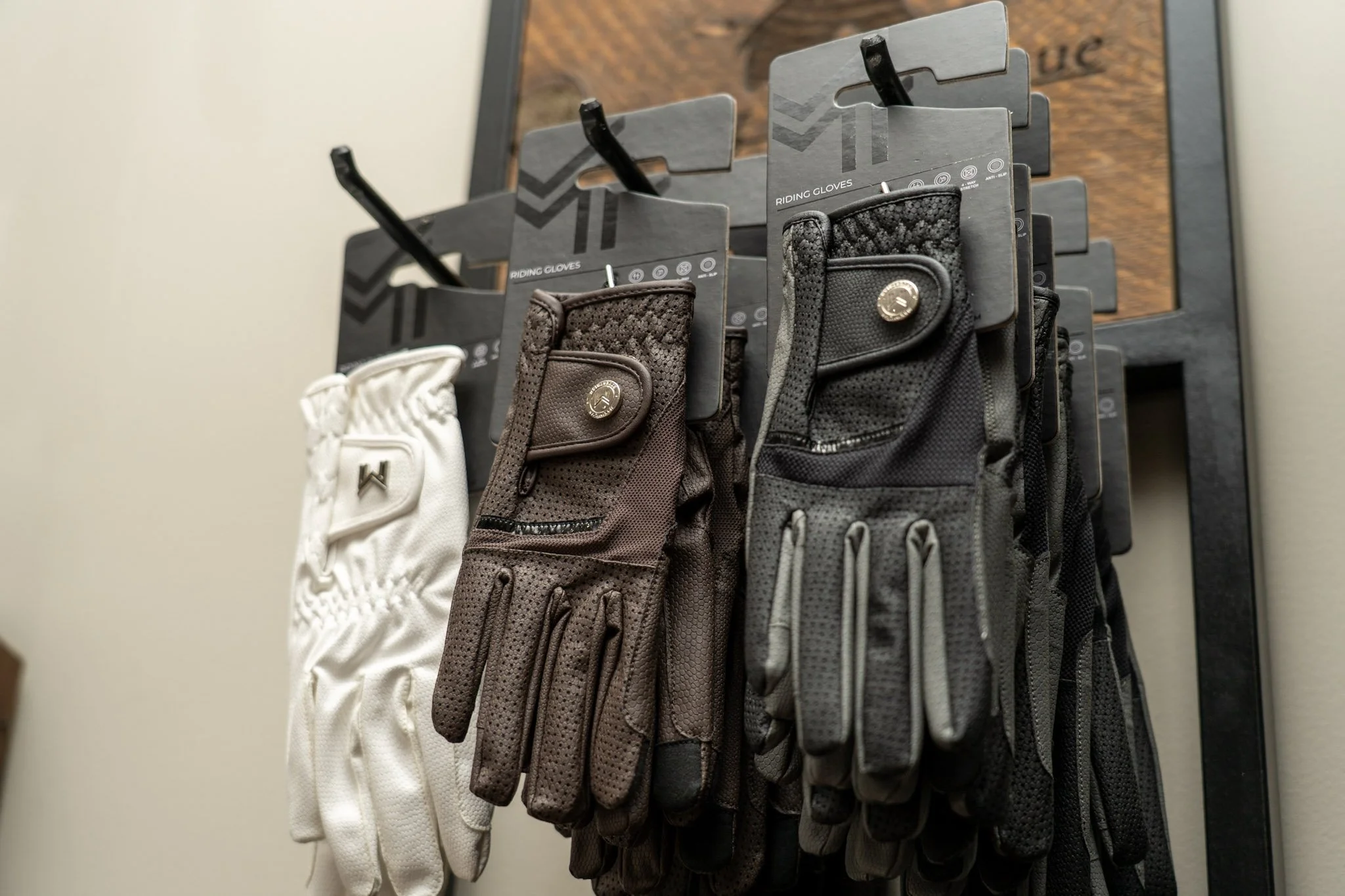 Display of cycling gloves in black, brown, gray, and white, hanging on hooks against a plaque with a wooden background.