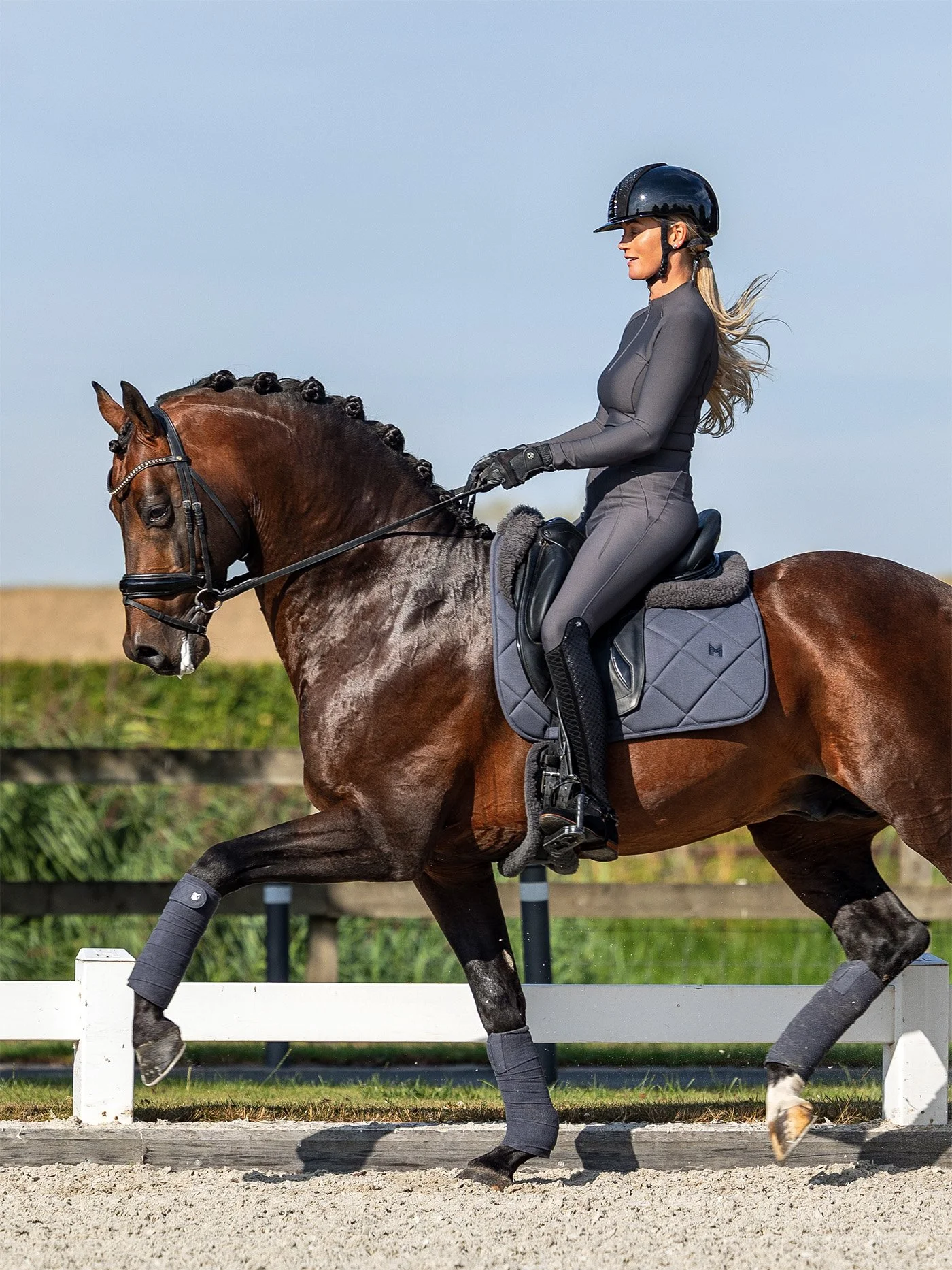 A woman wearing a helmet, riding gray horse with black mane, Galloping on outdoor riding arena with white fencing and green fields in background.