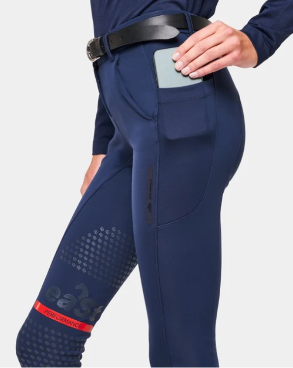 Person wearing blue athletic pants with a phone in the front pocket, and a hand reaching into the pocket. The pants are fitted and have a performance logo with a red performance label on the thigh.
