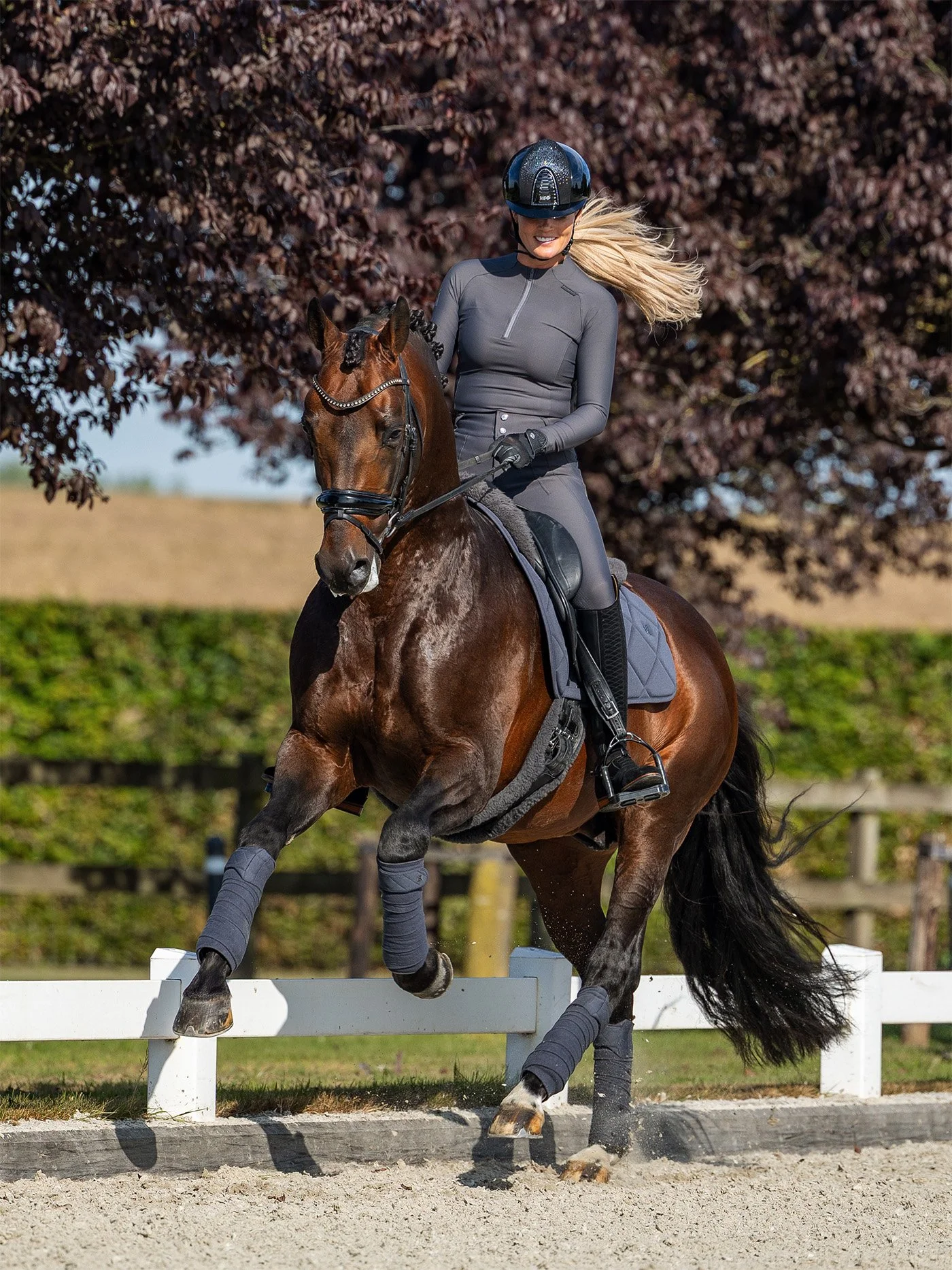 A woman riding a horse in a dressage arena during daytime, with autumn trees in the background.