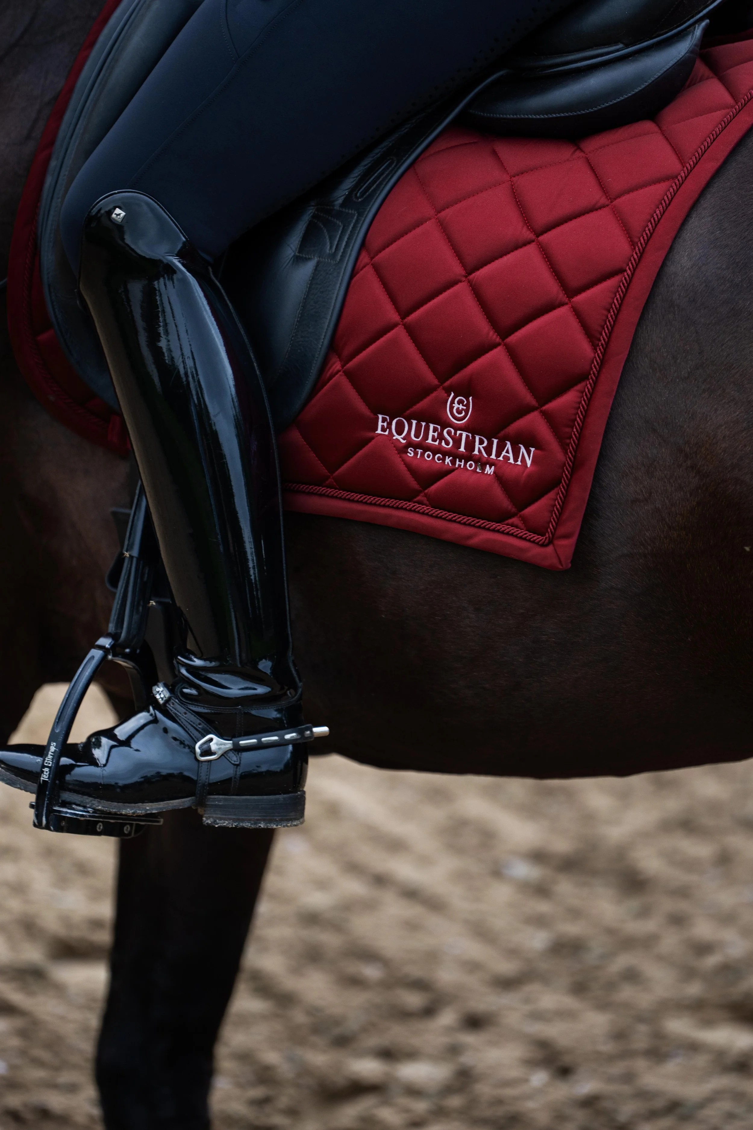 Close-up of a horse's saddle and riding boots on the horse's side, with a red quilted saddle pad embroidered with the words "EQUESTRIAN STOCKHOLM."