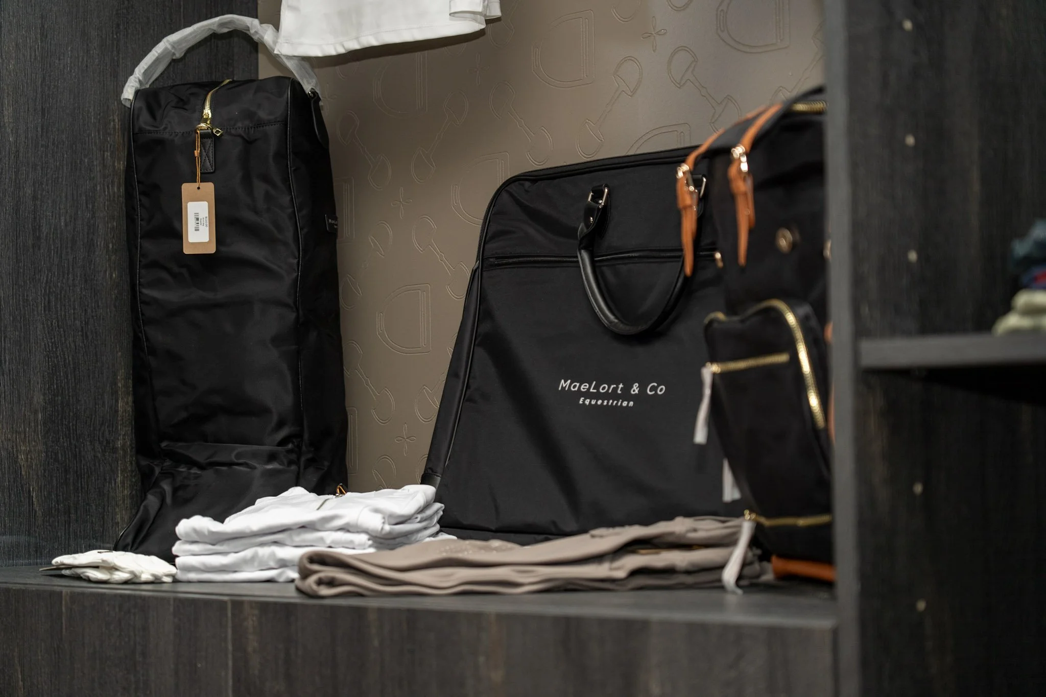 Display of folded white and beige clothes, black bags, and a folded black bag with orange straps on a dark shelf in a retail store.