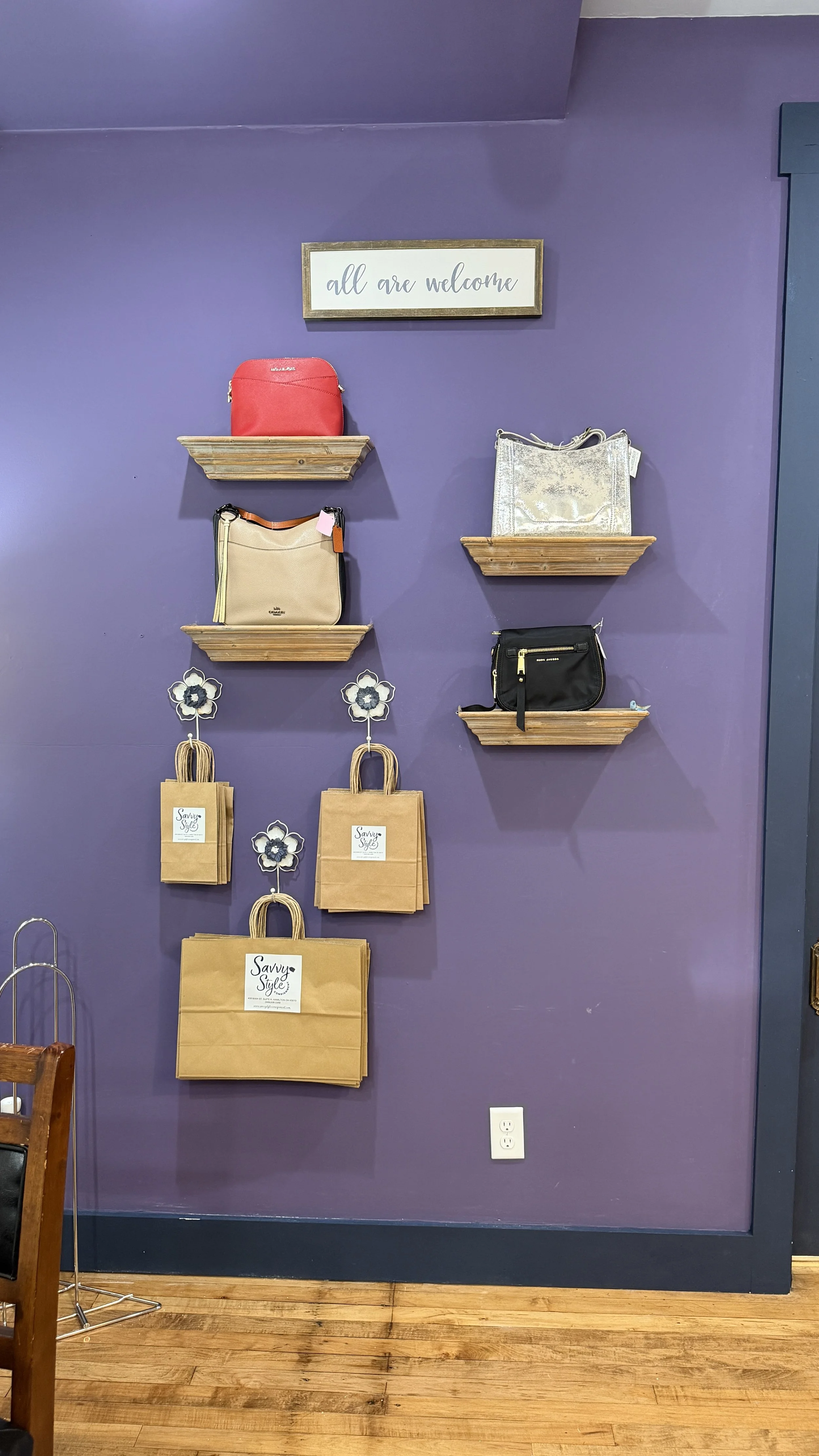 Display of handbags and gift bags on a purple wall with a sign that says 'all are welcome'