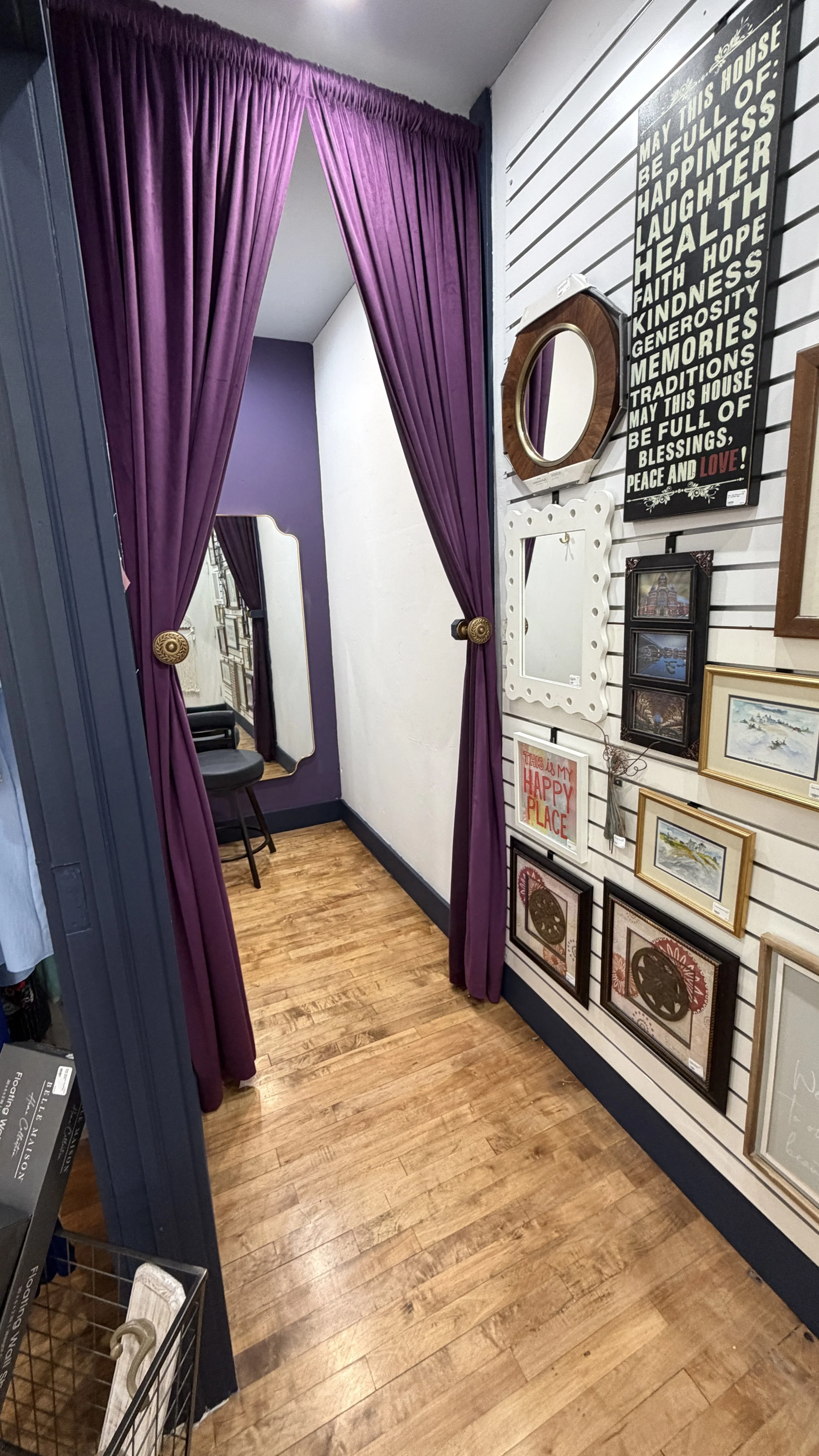 A fitting room with purple curtains and a mirror, surrounded by framed artwork and a sign with inspirational words on a white paneled wall.