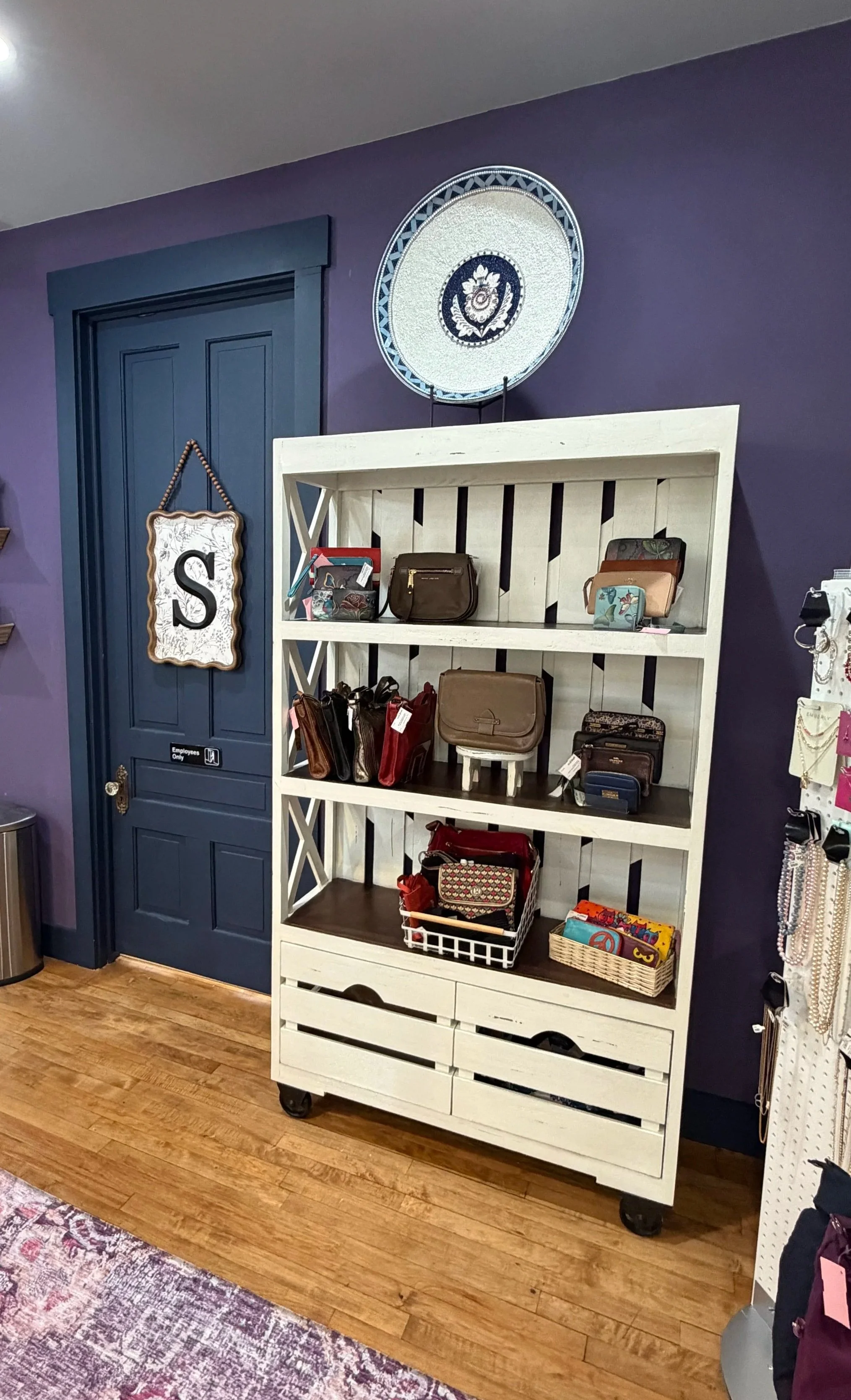 Retail display shelf with handbags, wallets, and accessories in a store, with a purple wall in the background and a blue door.