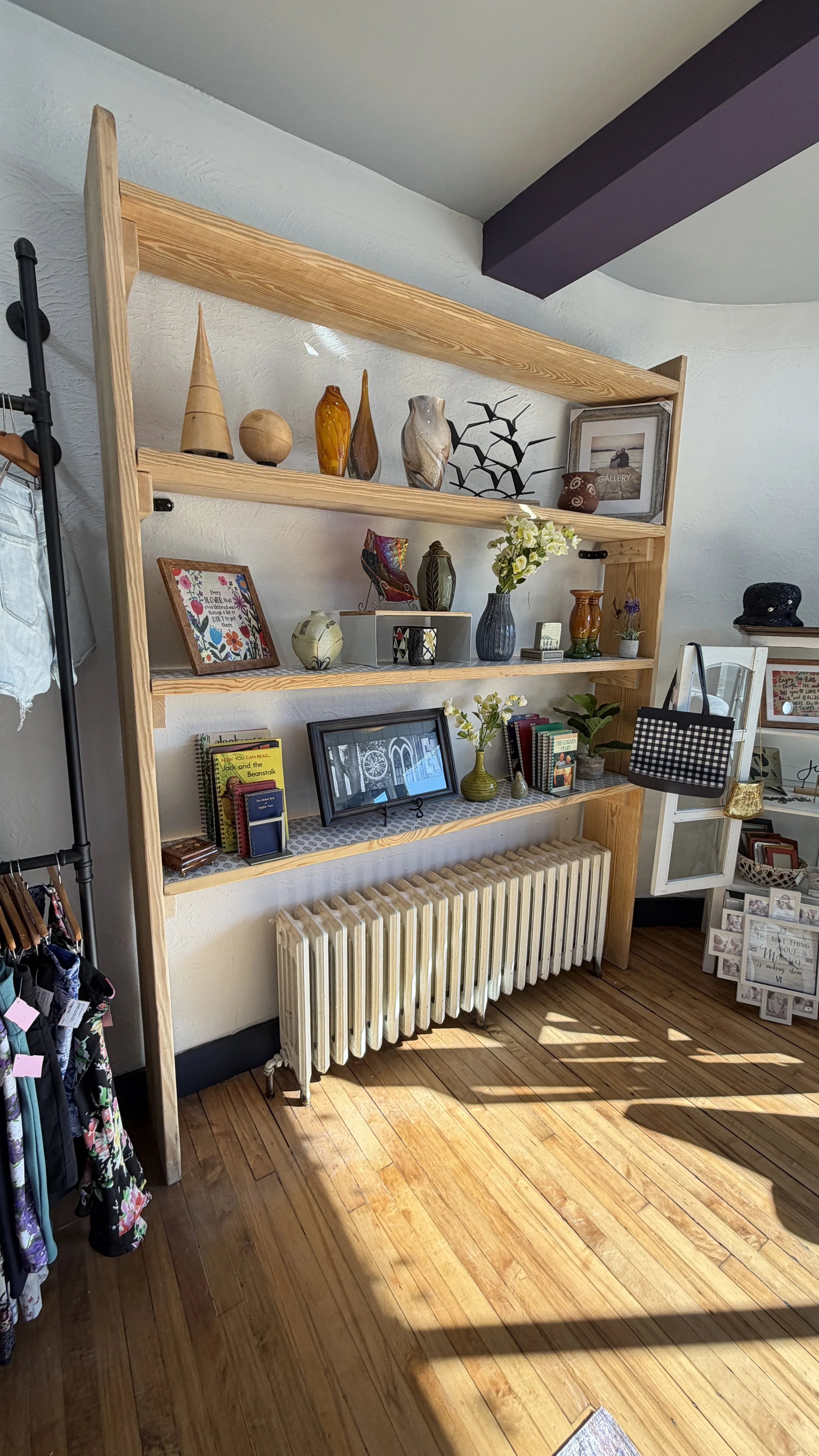 Wooden shelving unit displaying various decorative vases, framed pictures, and books in a cozy room with wooden flooring and a radiator underneath.