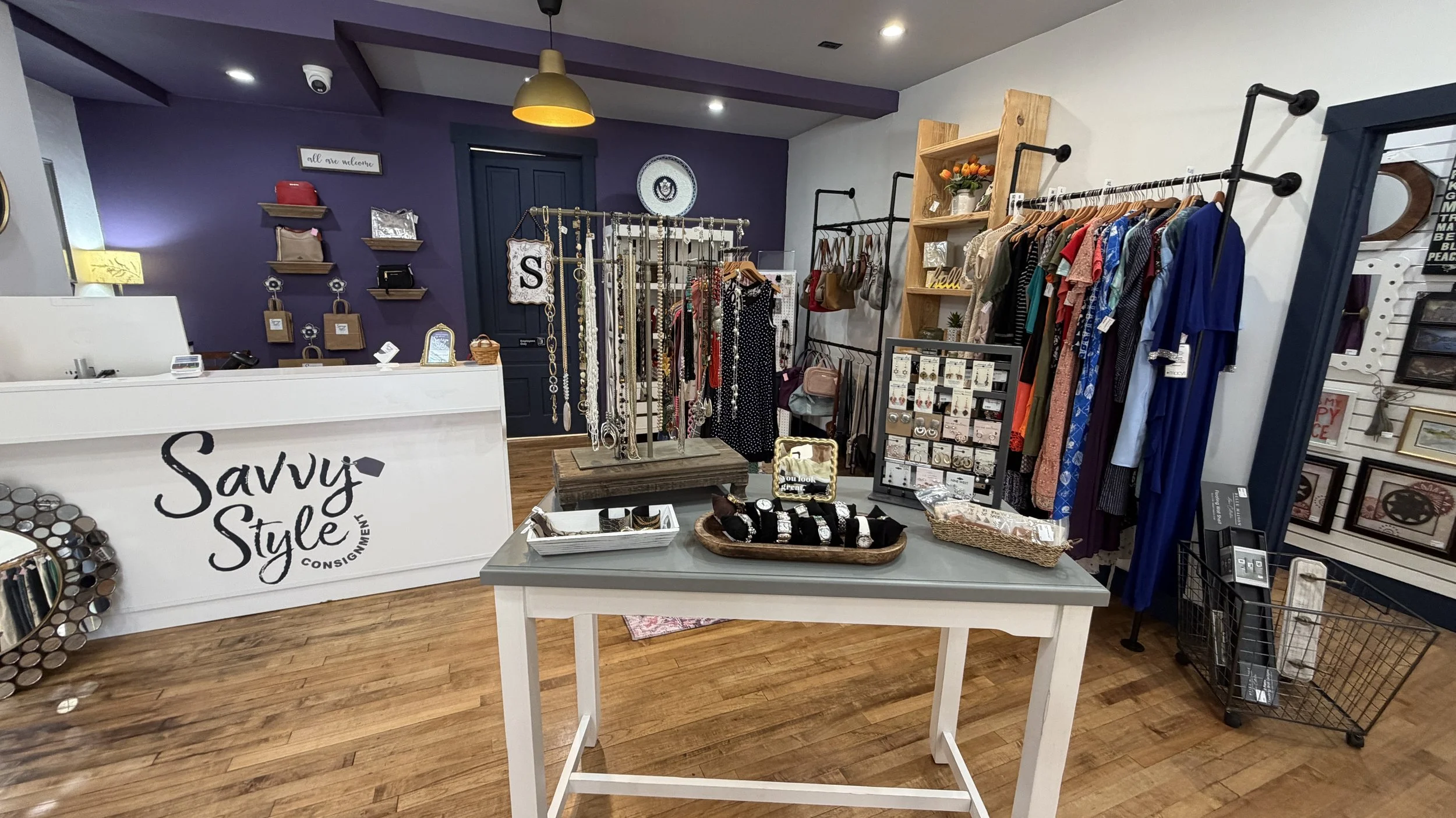 Interior of a boutique shop with a white checkout counter that has 'Savvy Style' written on it, racks of clothing, jewelry, and accessories, framed art, and a purple accent wall.