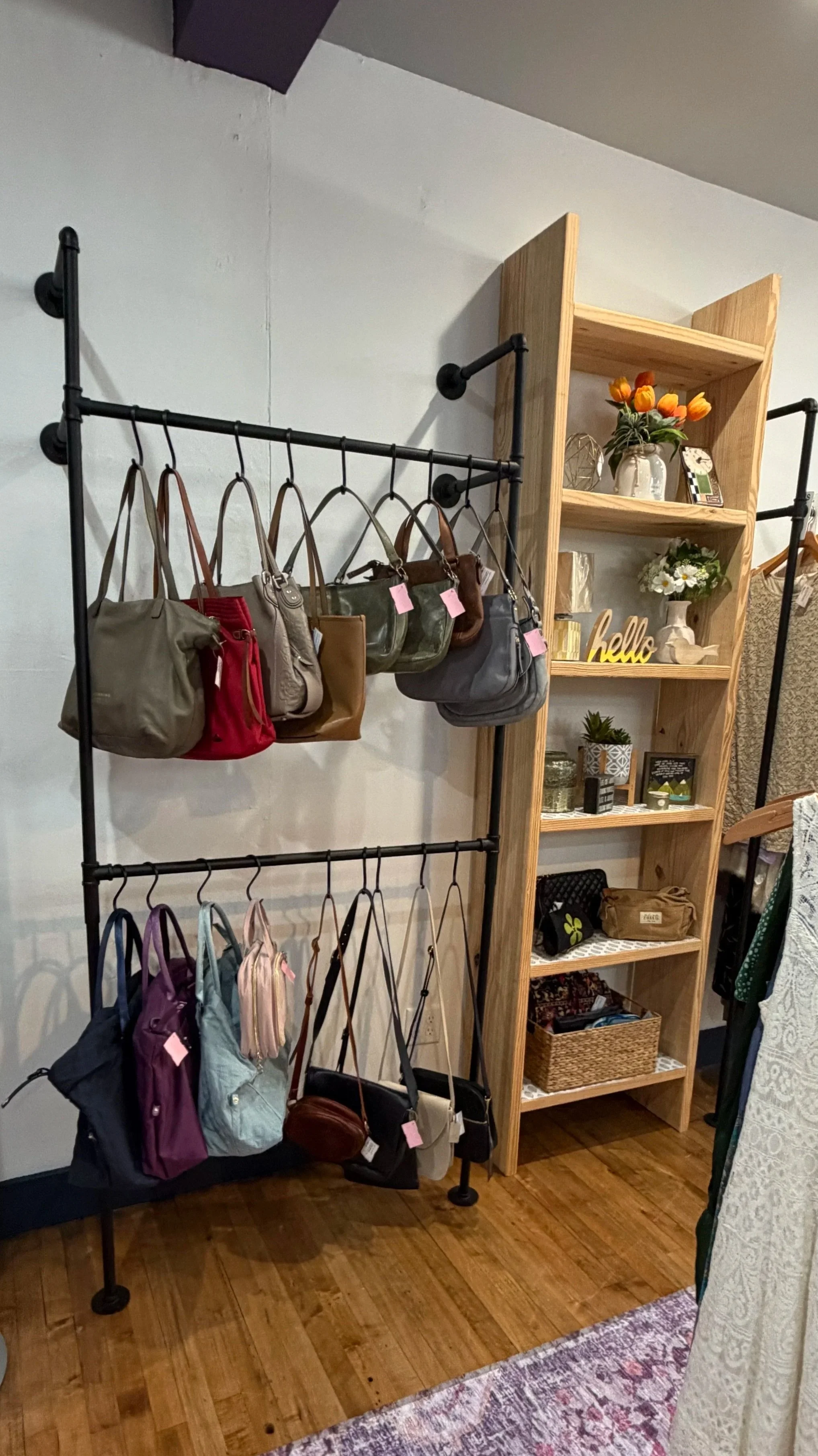 Display of handbags hanging on a black metal rack next to a wooden shelf with decorative items, flowers, and baskets in a boutique store.