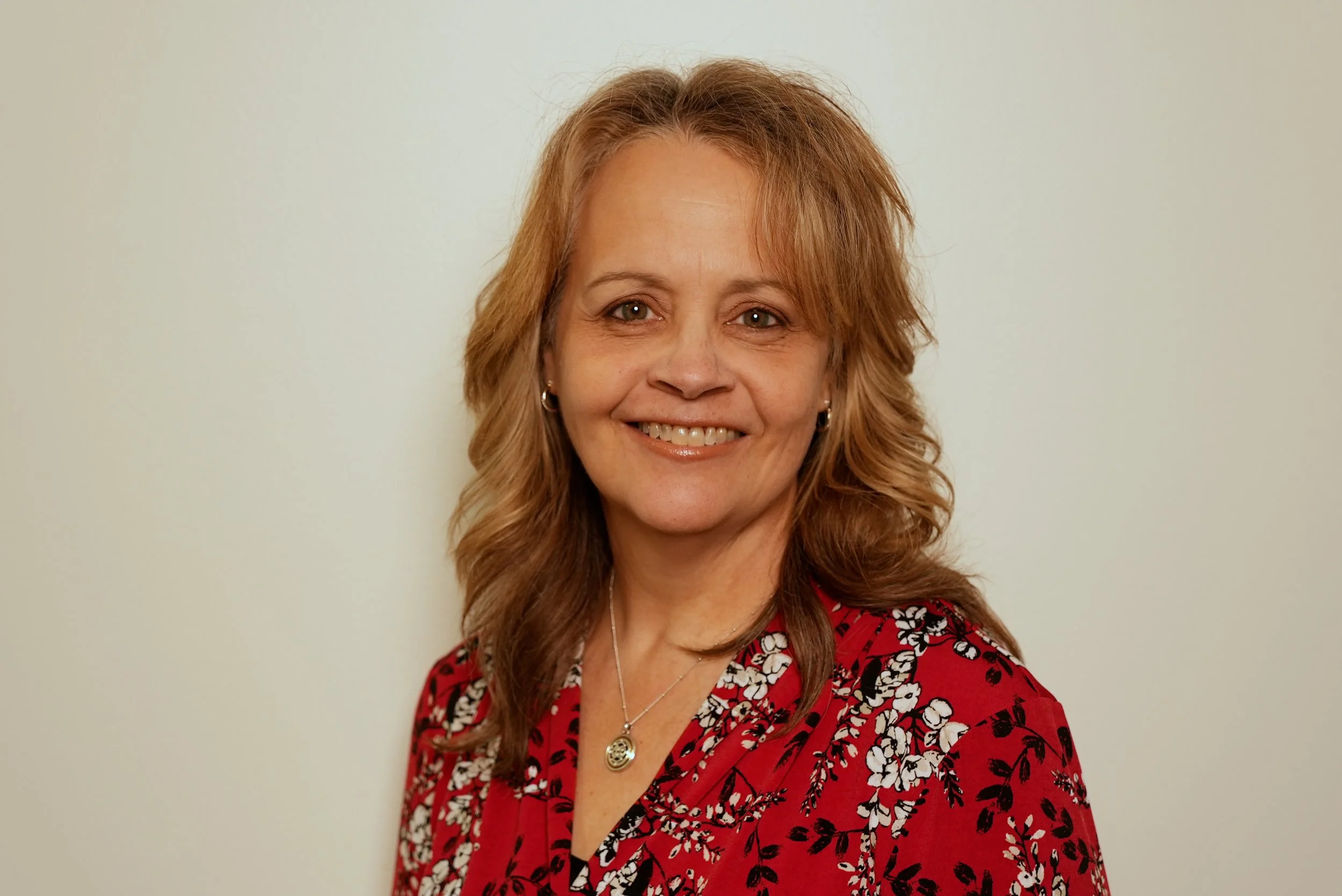 A smiling woman with shoulder-length wavy light brown hair, wearing a red dress with a white and black floral pattern, a silver necklace with a round pendant, and small earrings, standing against a plain off-white wall.
