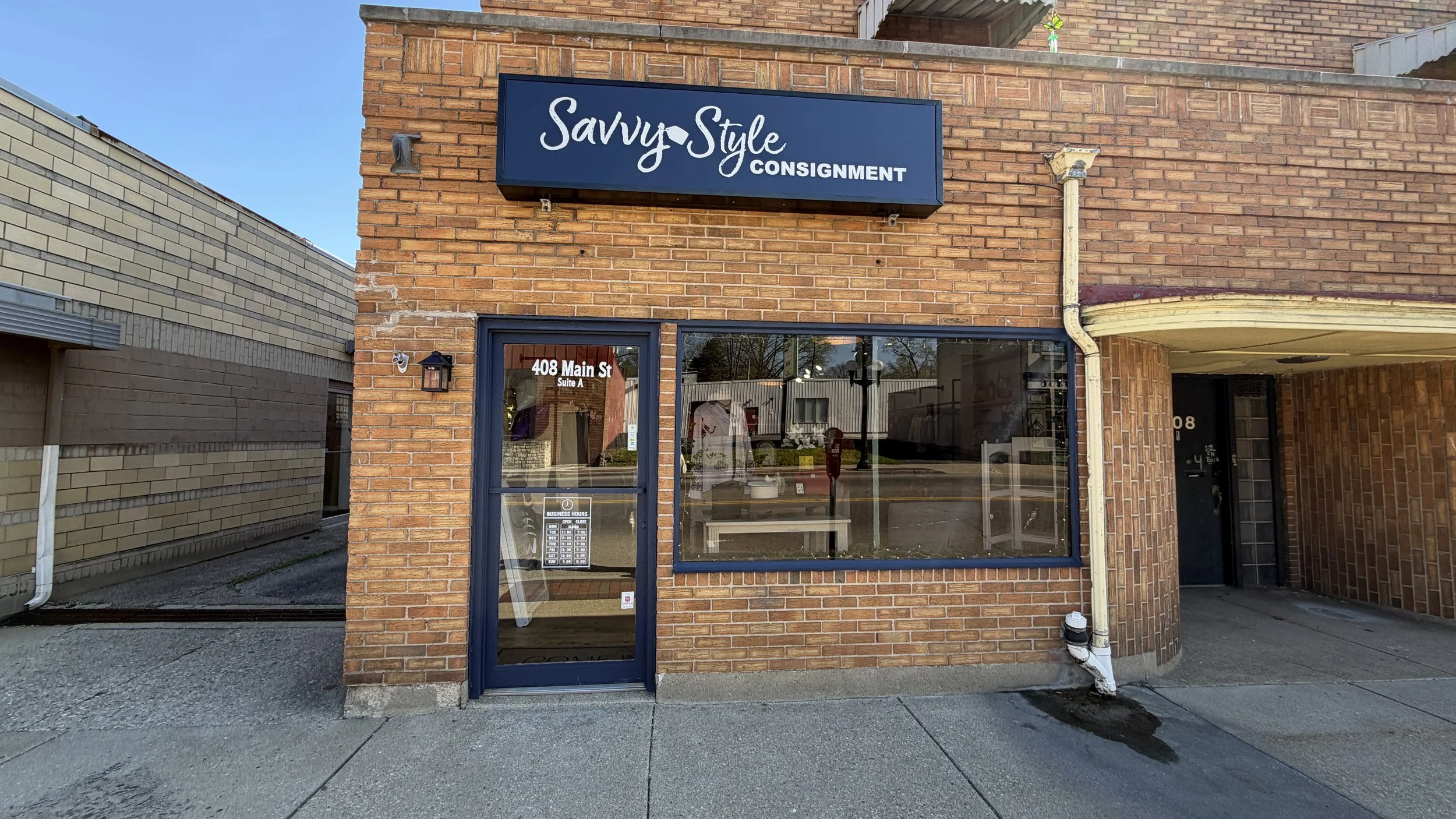 Brick storefront with a sign that reads 'Savvy Style CONSIGNMENT'. The entrance has a glass door and large window, with the address '408 Main St' on the door.