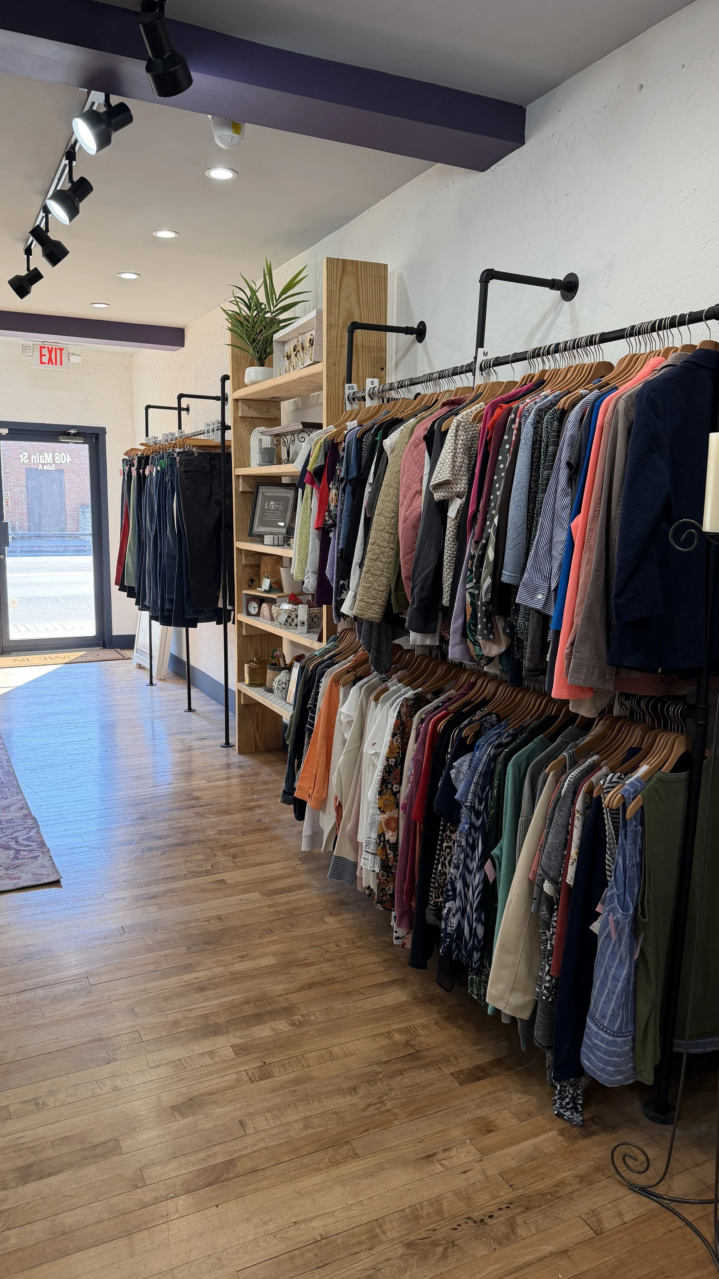Inside a clothing thrift store with rack of assorted clothes on both sides, a wooden shelf with decorative items and framed pictures, and a glass door at the front letting in natural light, with wooden flooring.