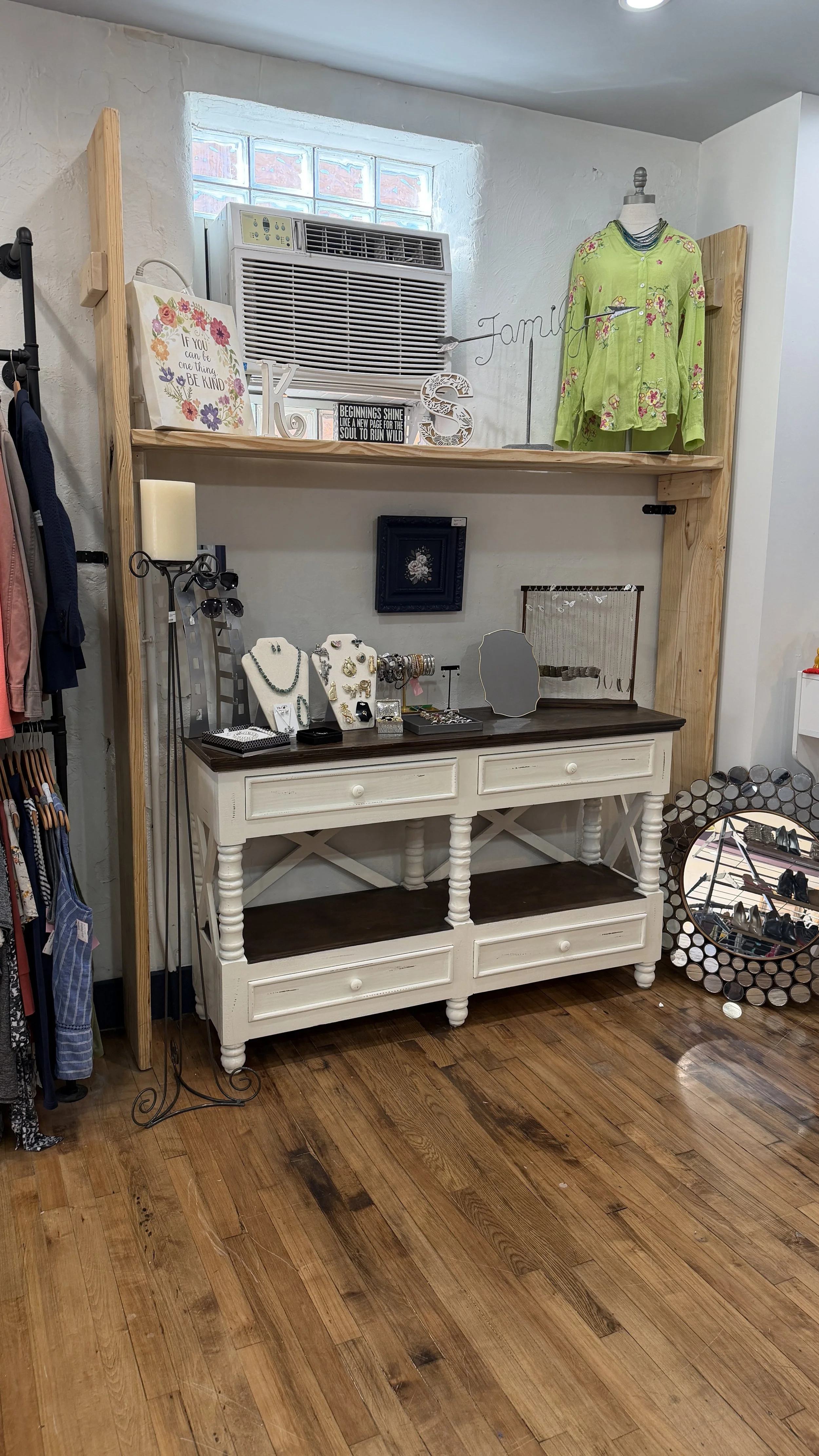 A retail display with jewelry, accessories, and clothing items on a white and dark wooden dresser inside a store. The background includes a small window, an air conditioning unit, and wall decor.