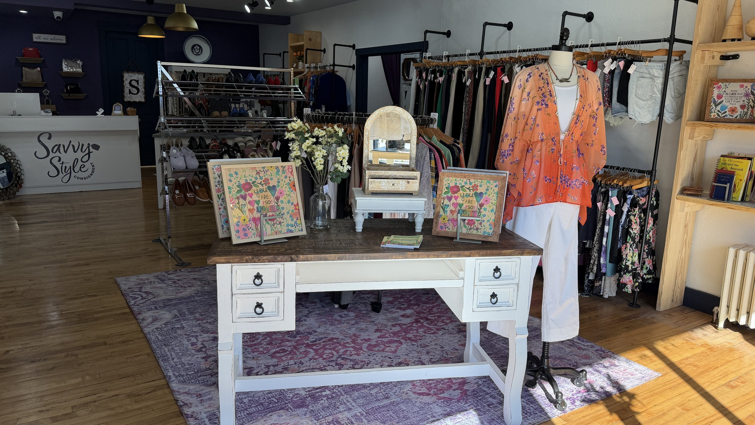 A boutique clothing store with a white vintage desk in the foreground, topped with decorative items including framed art, a glass vase with white flowers, and a mirror. Behind the desk, there are racks of clothing, a mannequin dressed in orange and white, and shelves filled with books and decorative objects. The store has hardwood floors and a cozy, eclectic decor.