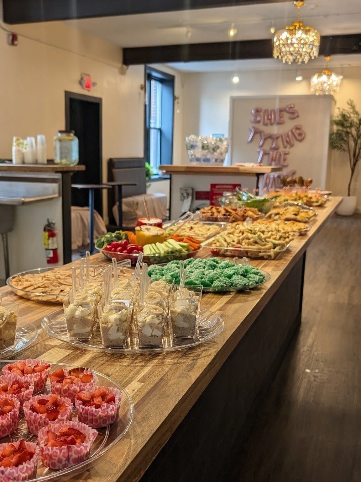 A buffet table with various desserts and snacks, including cupcakes, fruit, and candy, at an indoor event space decorated with chandeliers and a "she's tying the knot" sign in the background.