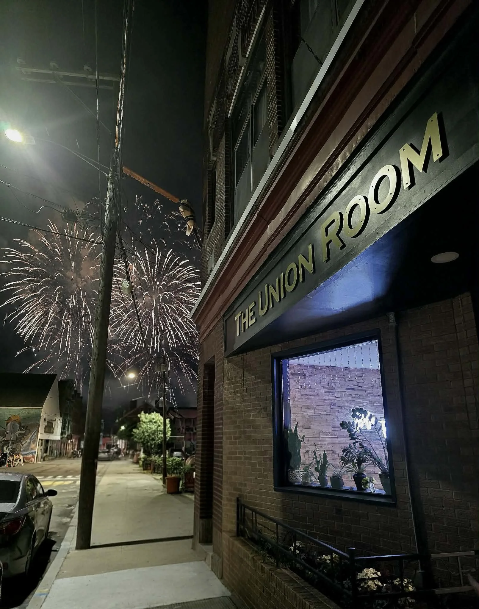 Nighttime street scene with fireworks in the sky, a brick building with a sign reading 'The Union Room,' plants on a window ledge, streetlights, parked cars, and utility poles with wires.