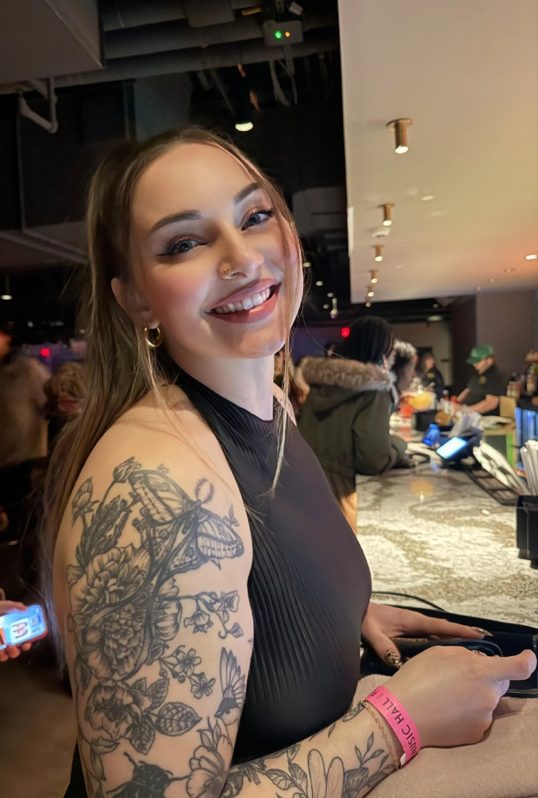 A young woman with tattoos on her arm, smiling at the camera in a busy indoor setting, possibly a bar or restaurant.