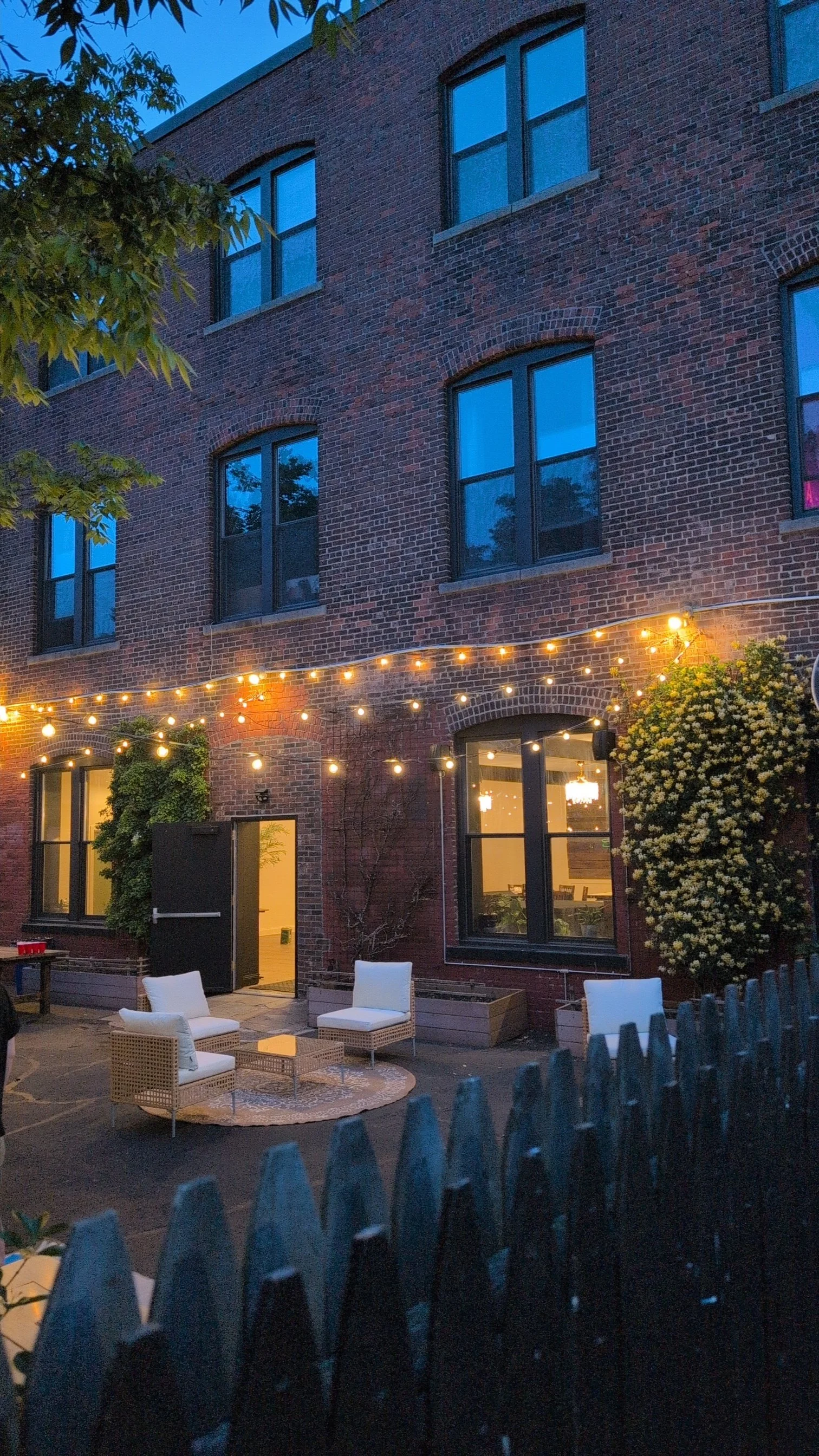 A brick apartment building with multiple tall windows, some illuminated from inside. Outdoor string lights hang across a patio area with white chairs and a small round table, surrounded by a picket fence. There are trees and bushes, and the scene is captured during dusk or evening.