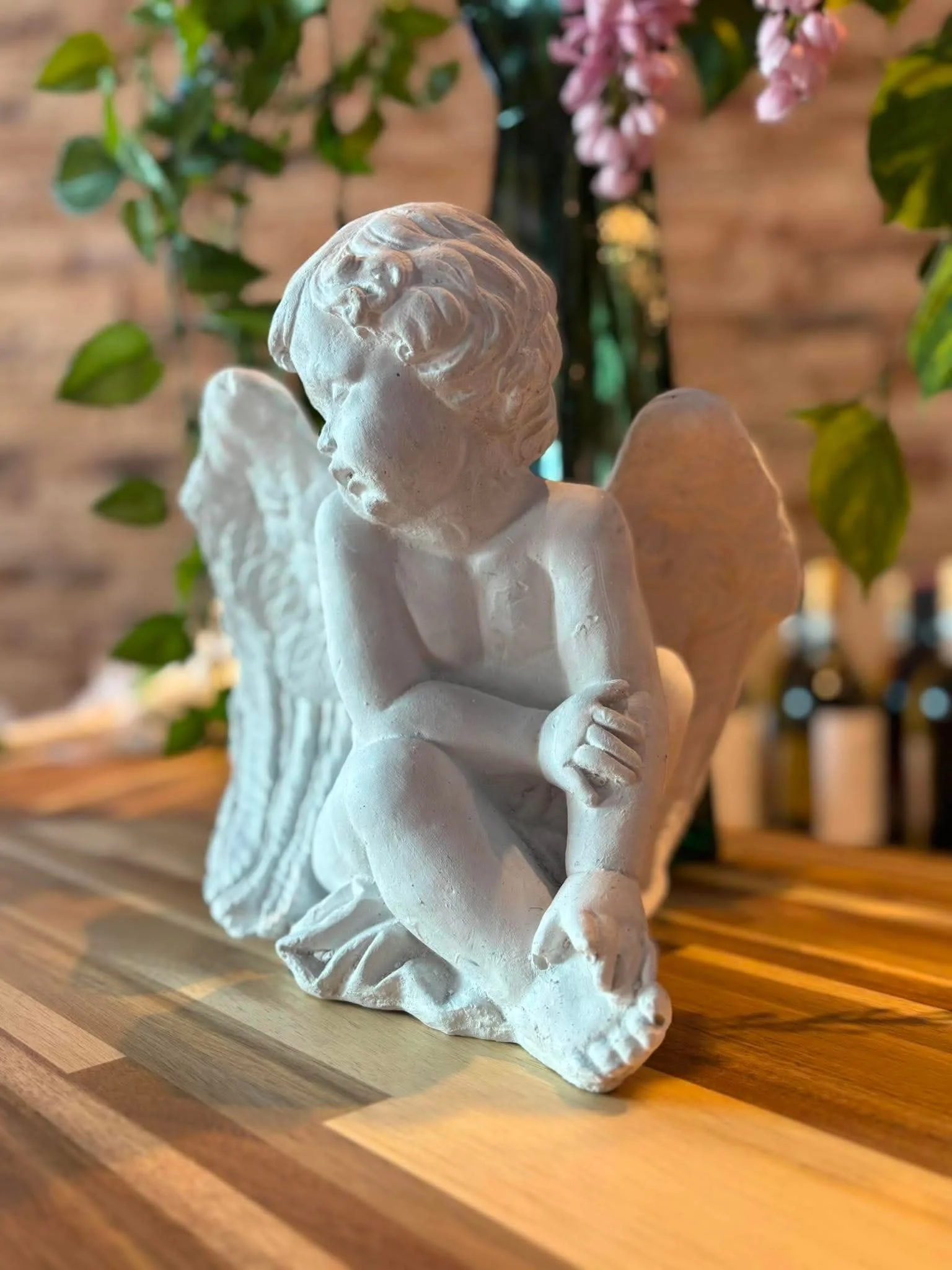 A white ceramic angel figurine with closed eyes and clasped hands, with multicolored wood surface and plants and pink flowers in the background.