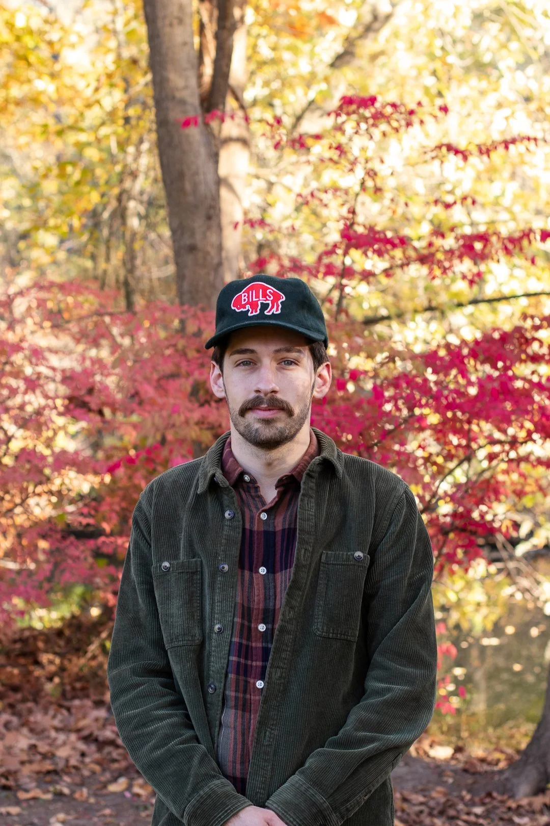A man with a mustache and beard stands outdoors during autumn, wearing a black cap with a red and white Buffalo Bills logo, a dark green jacket, and a plaid shirt, with colorful fall foliage in the background.