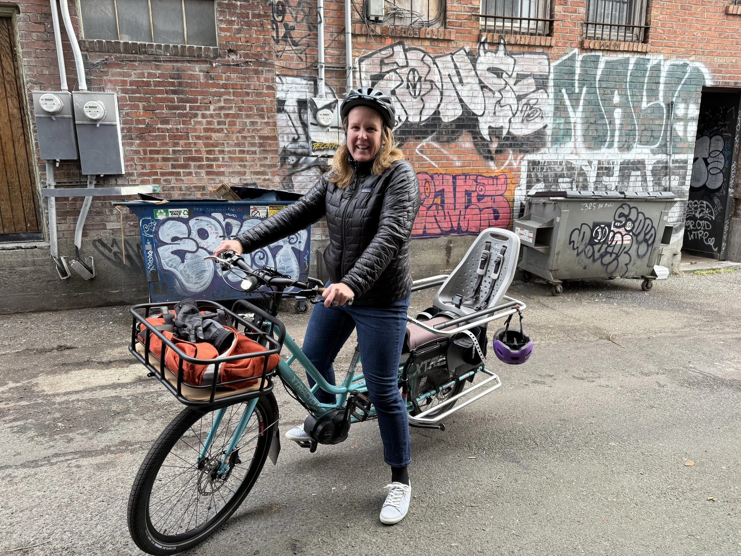 Woman riding a teal electric bike with a child seat, wearing a black jacket and helmet, on a city street with graffiti on brick walls and two trash bins.