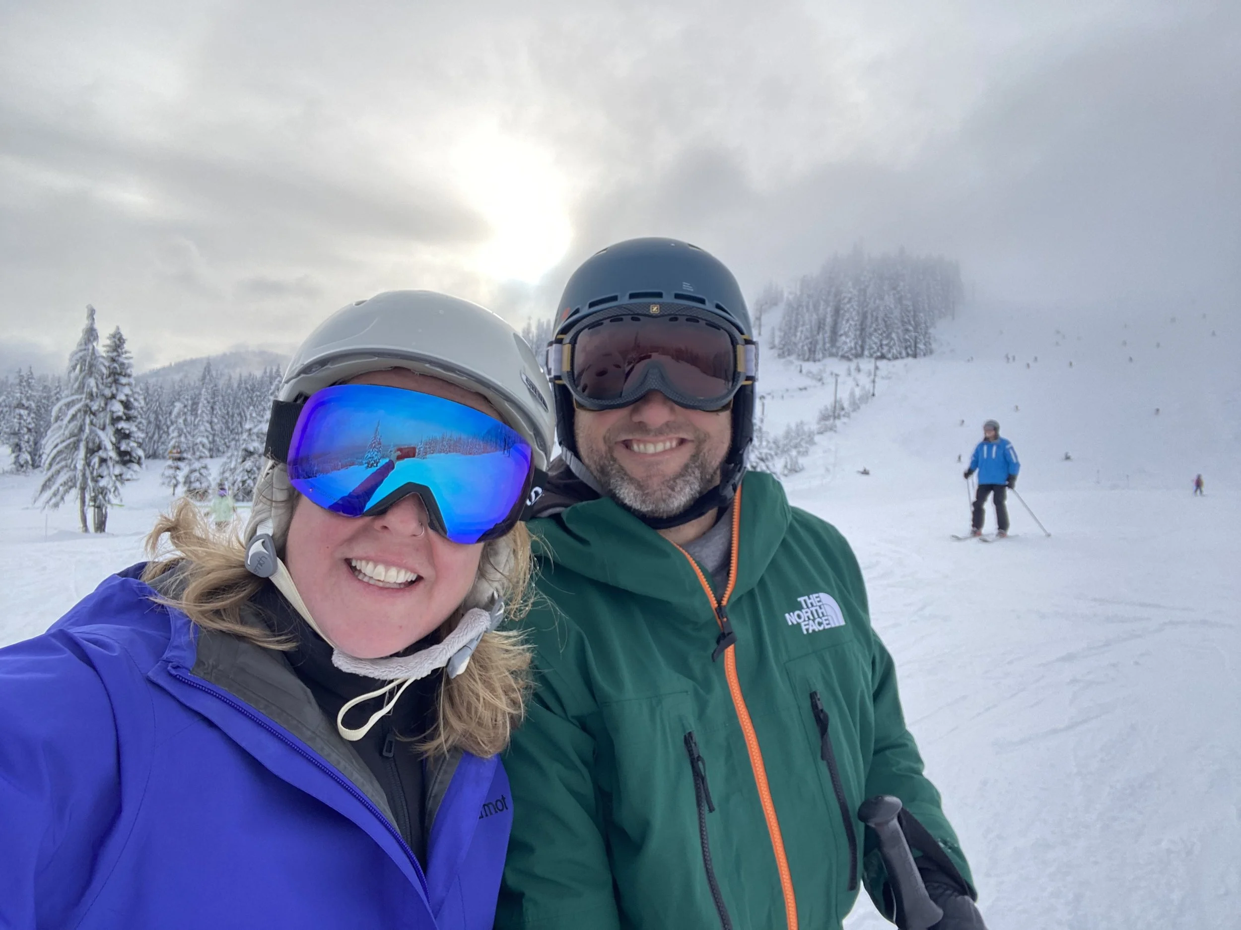 A smiling couple wearing ski helmets and goggles taking a selfie on a snowy mountain with skiers in the background.