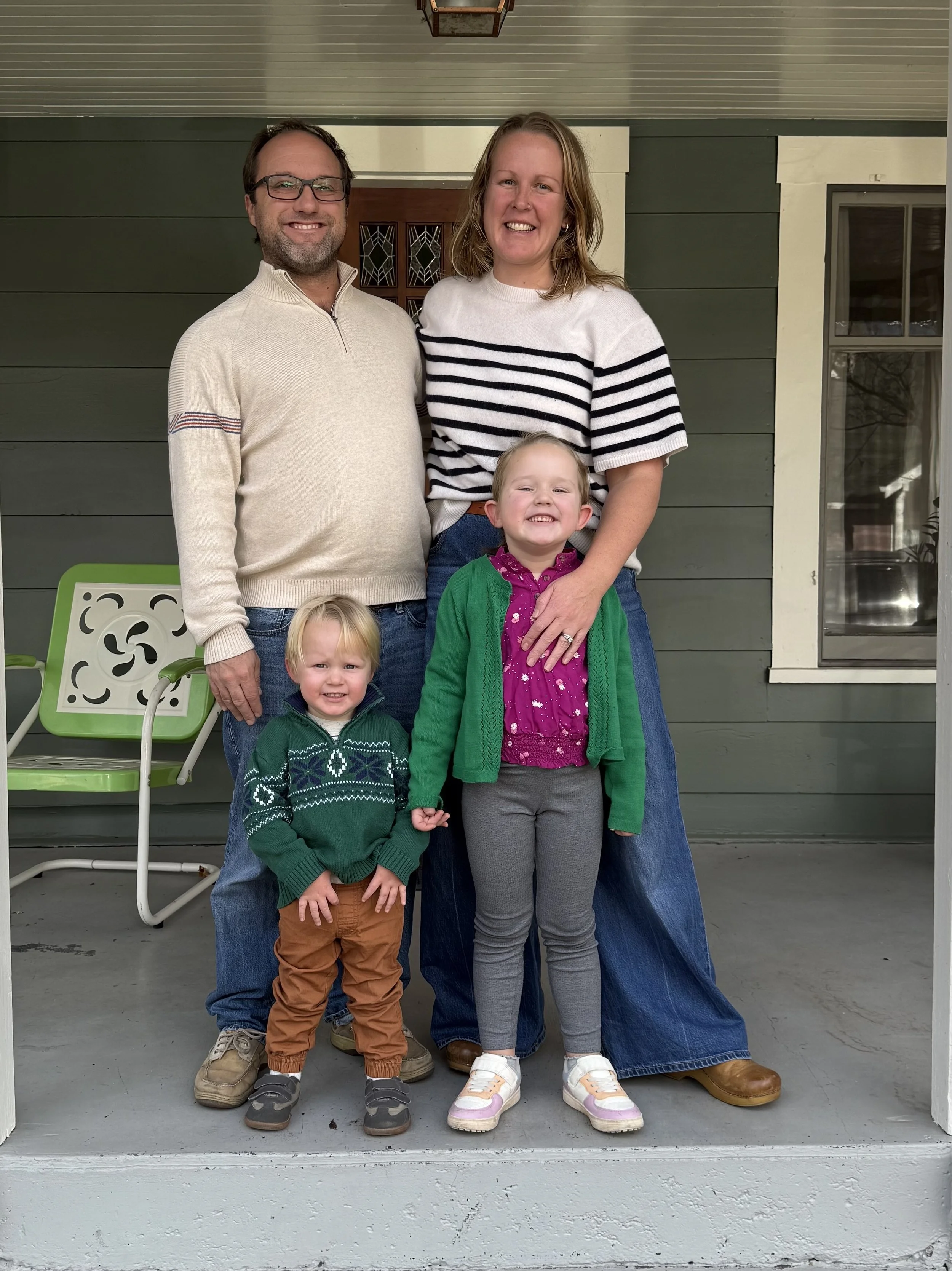 A family of four standing on a porch in front of a house, smiling at the camera. Two adults, a man and a woman, and two young girls. The man has glasses and a beard, wearing a light-colored sweater and jeans. The woman has shoulder-length hair, wearing a striped shirt and jeans. One girl is wearing a purple shirt with a green cardigan and leggings, and the other girl is wearing a green sweater with brown pants. There is a green and white chair on the porch and the house has green siding with a window.