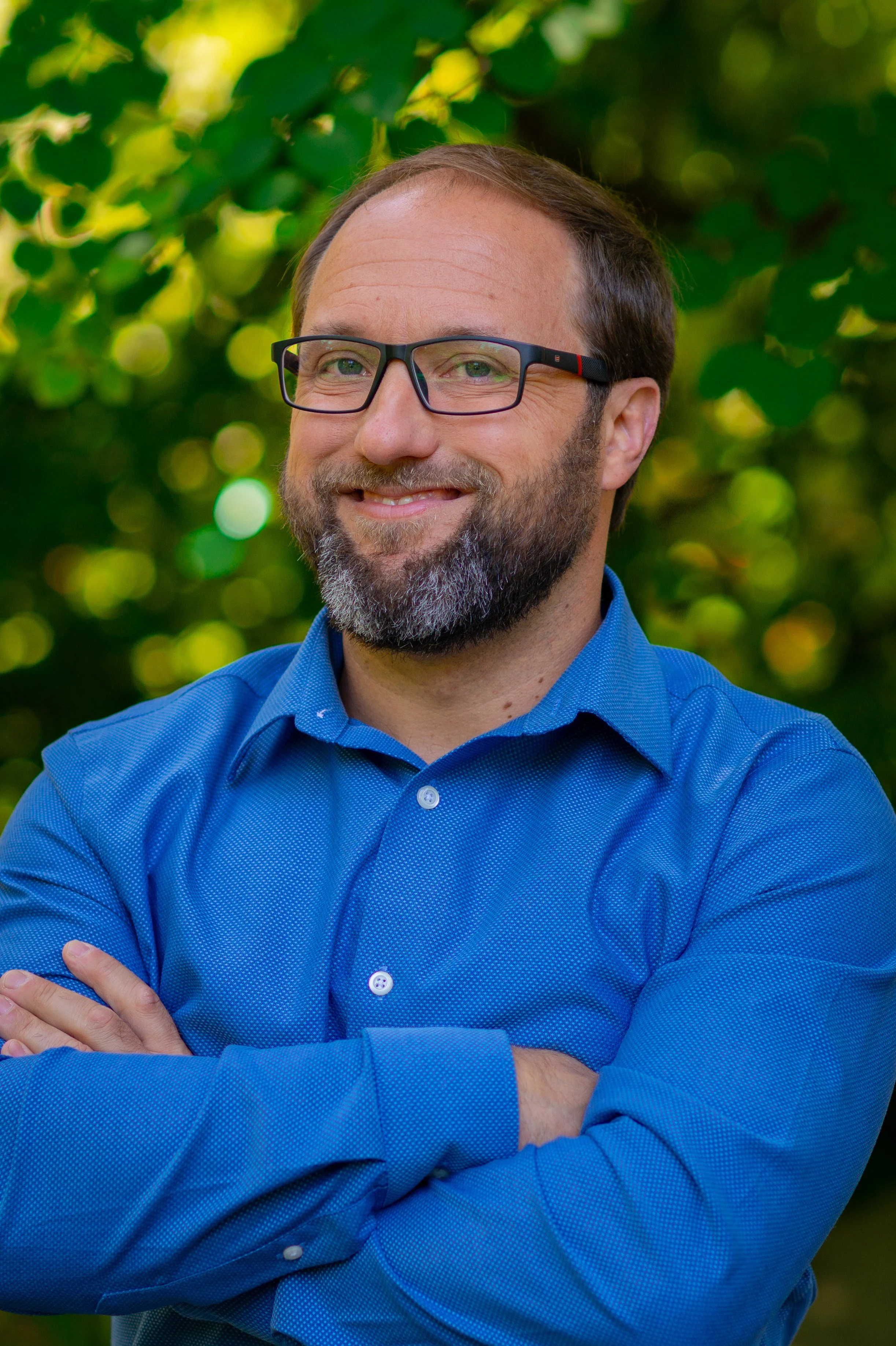 A man with glasses and a beard, wearing a blue button-down shirt, smiling with his arms crossed outdoors with greenery in the background.