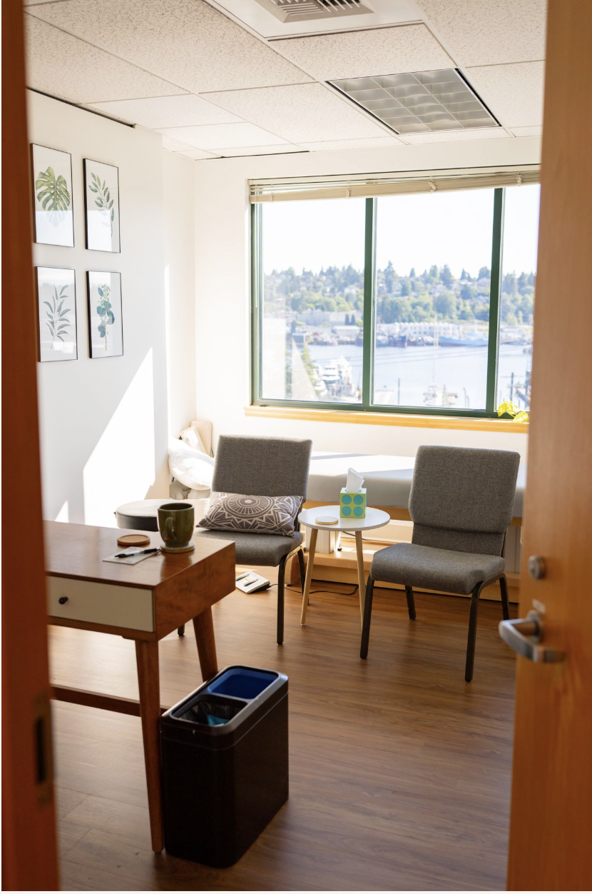 doorway into doctor's office. There is a window in the background overlooking seattle's mountains. two chairs are both positioned in front of exam table and facing a desk. The room is bright with natural light.