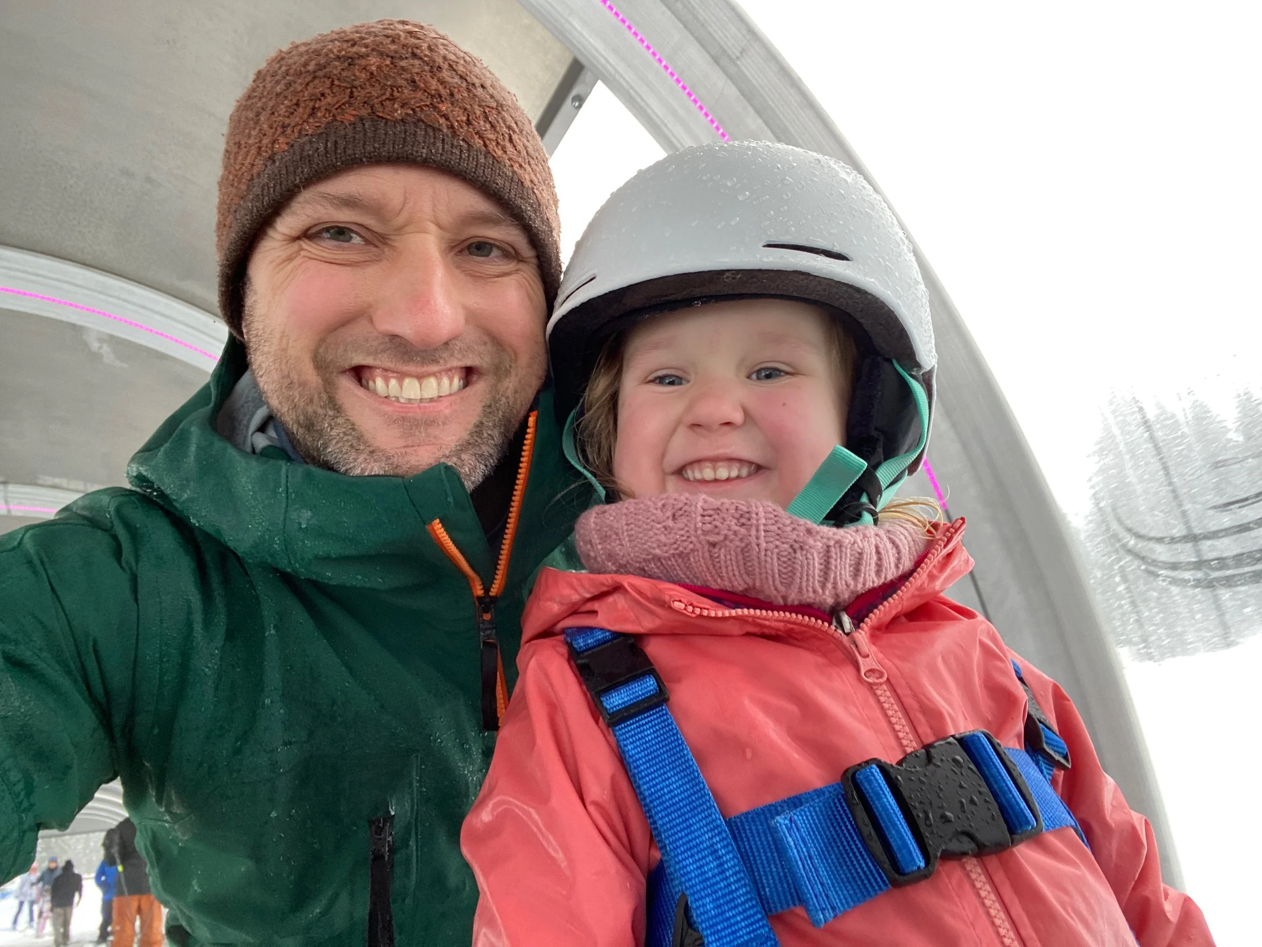 Man and young girl taking a selfie inside a snow-covered ski lift or gondola, both in ski gear, smiling