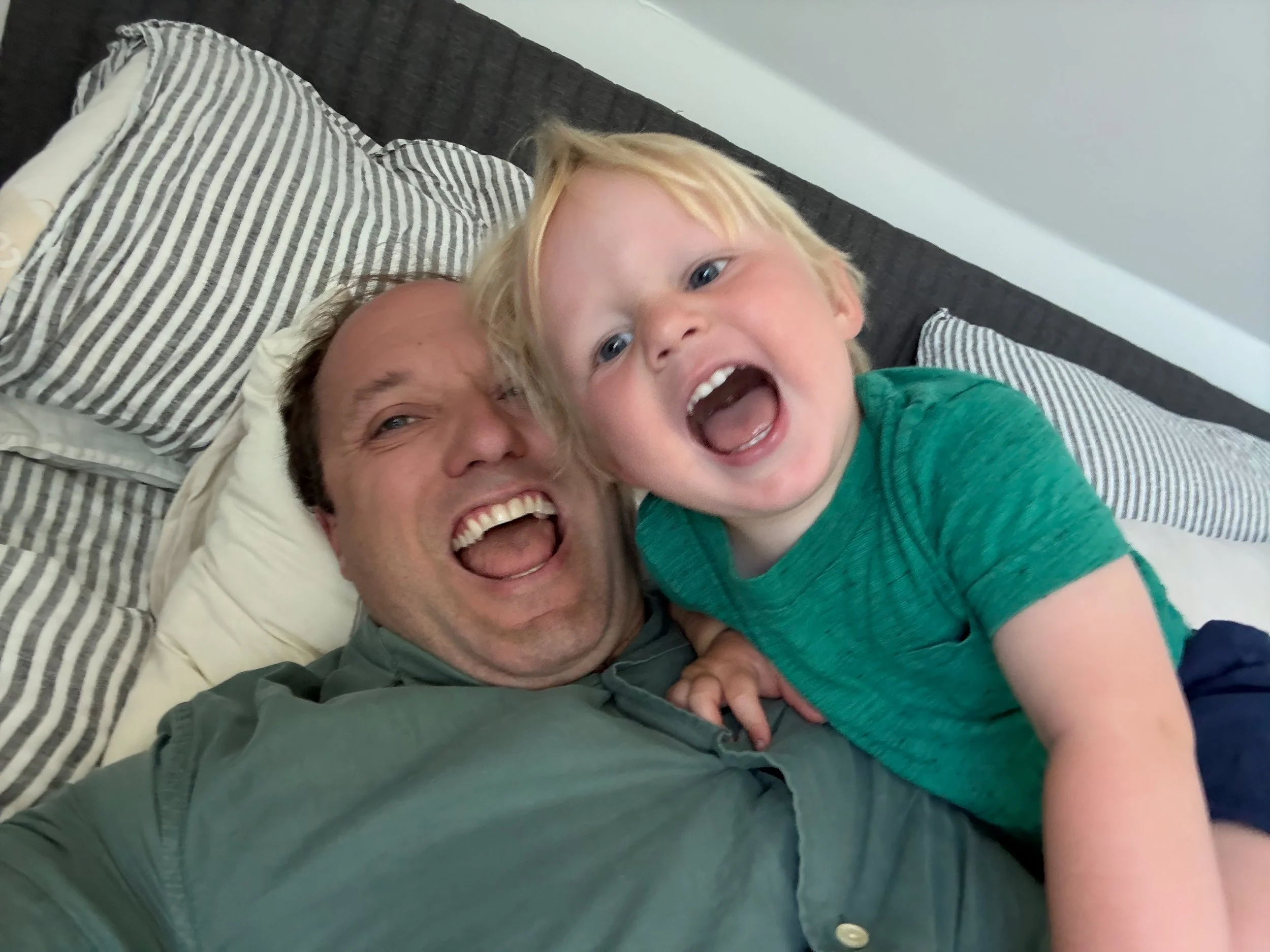 A man and a young boy on a bed, both smiling and laughing. The man has short brown hair and is wearing a dark green shirt, while the boy has blonde hair and is wearing a green t-shirt.
