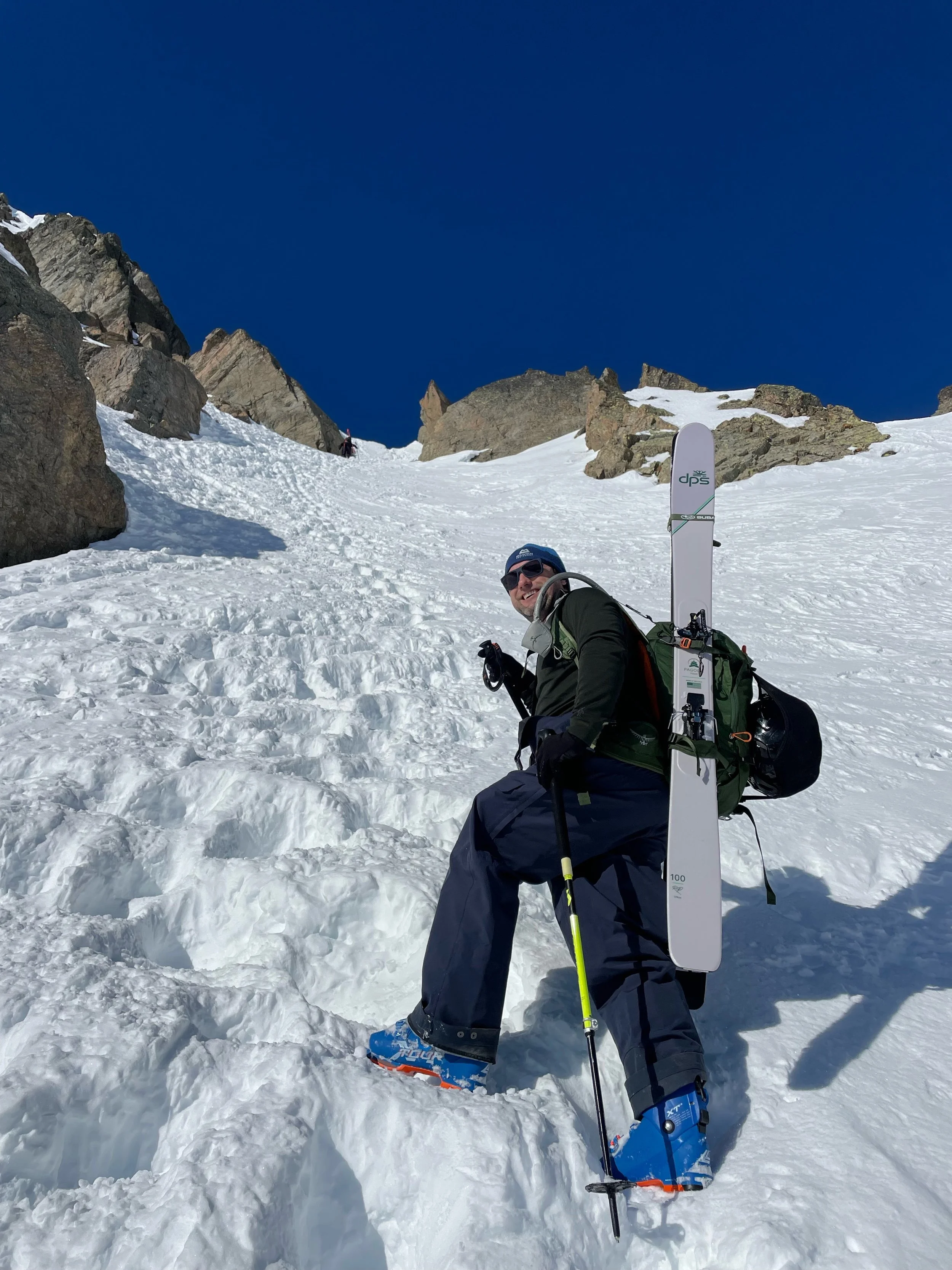 A man in snow gear climbing a snowy mountain slope with an ice axe, wearing crampons, sunglasses, a backpack, and holding skis on his back, under a clear blue sky.