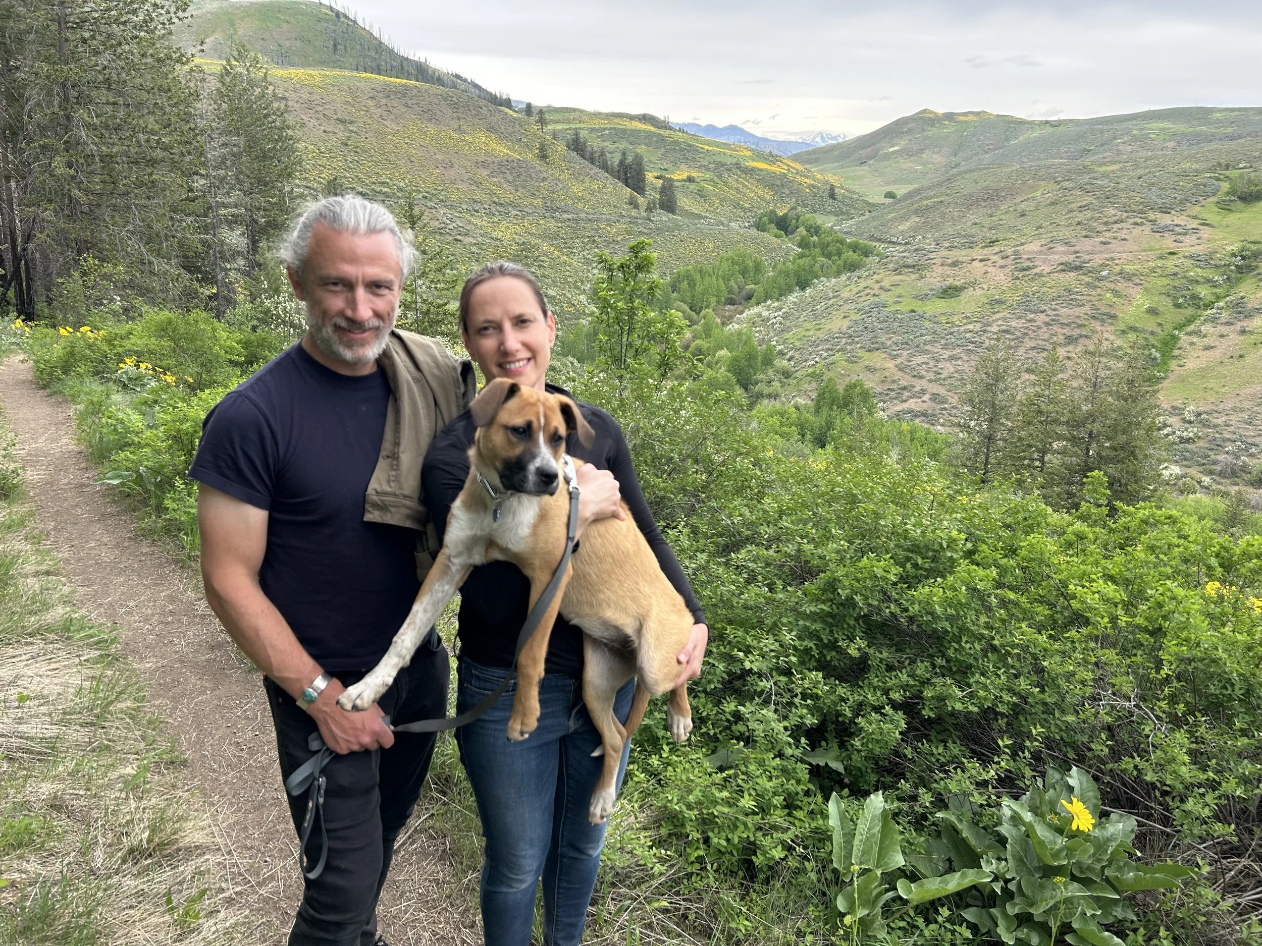 A man and woman standing on a trail in a lush, green valley with rolling hills and mountains in the background, holding a puppy between them.