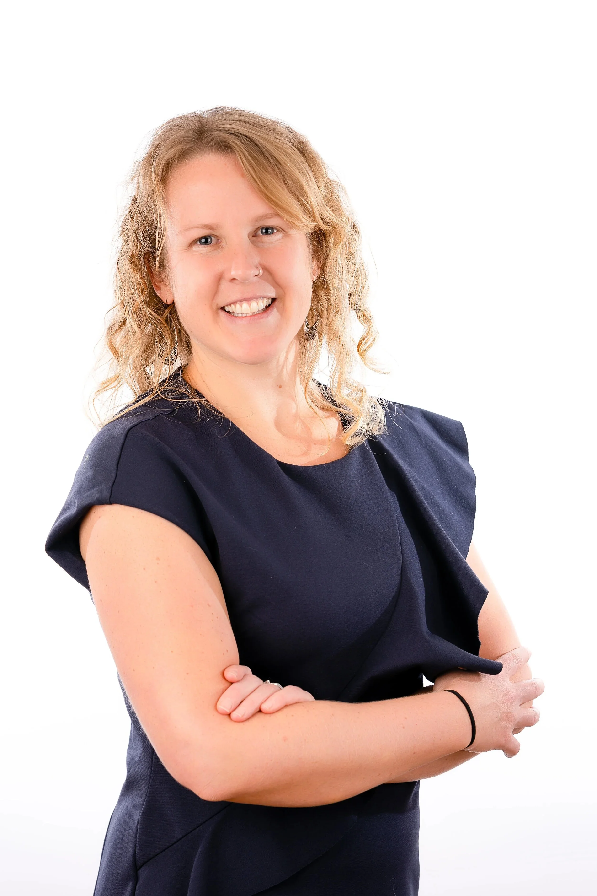 Portrait of a smiling woman with curly blonde hair wearing a dark blue dress with folded sleeves, standing with her arms crossed against a white background.