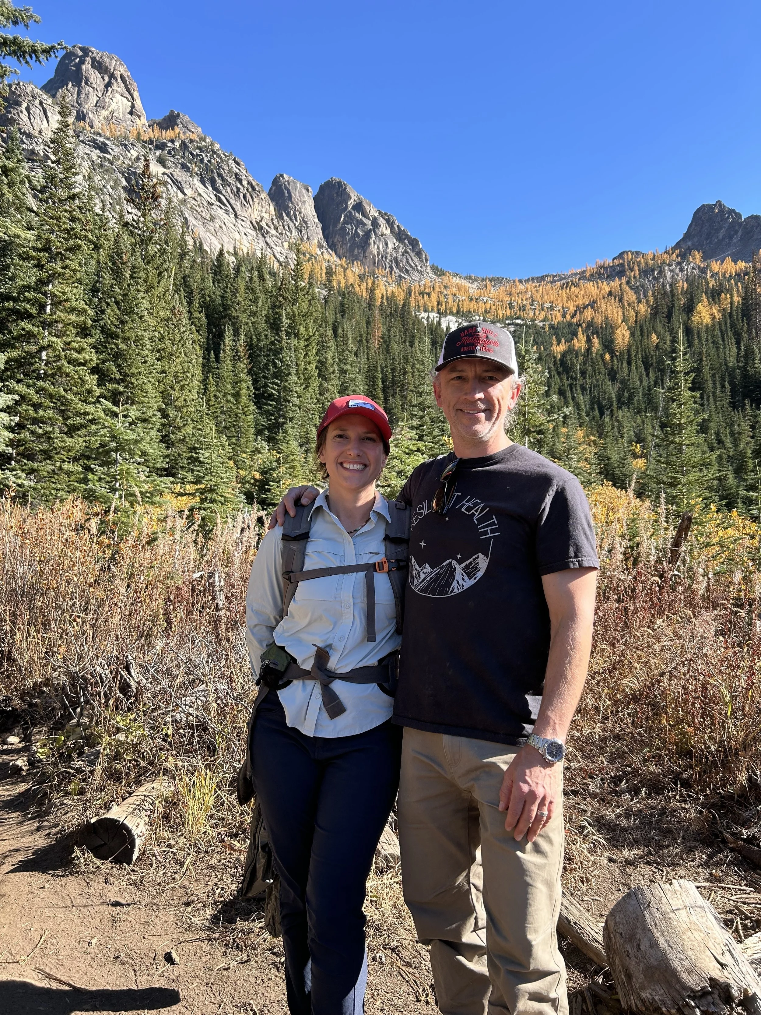 A smiling couple standing outdoors in a forested mountain area, with tall trees, rocky mountains, and a clear blue sky in the background, dressed for hiking.