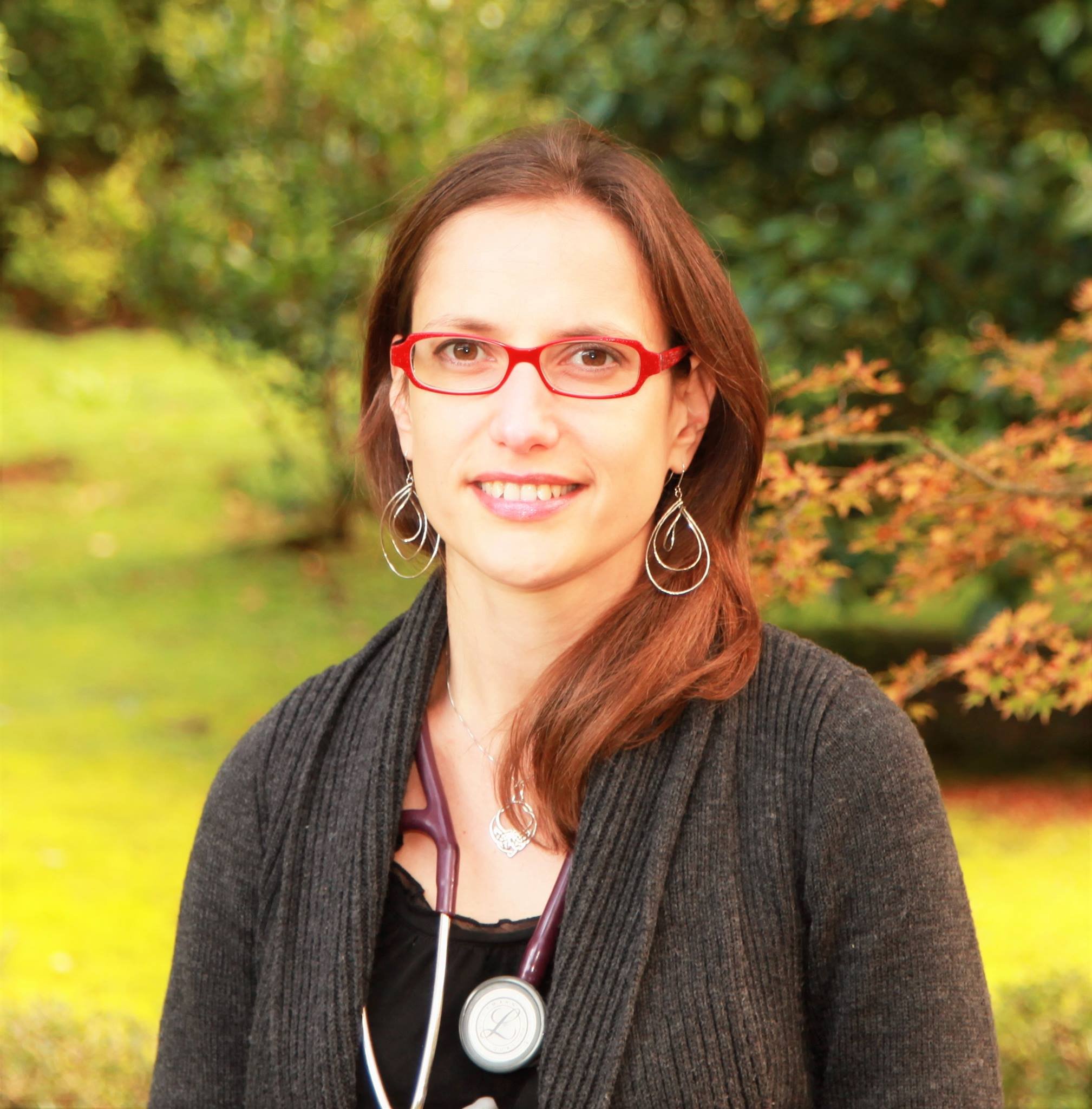 A woman with red hair and red glasses smiling outdoors with autumn leaves and a pond in the background.