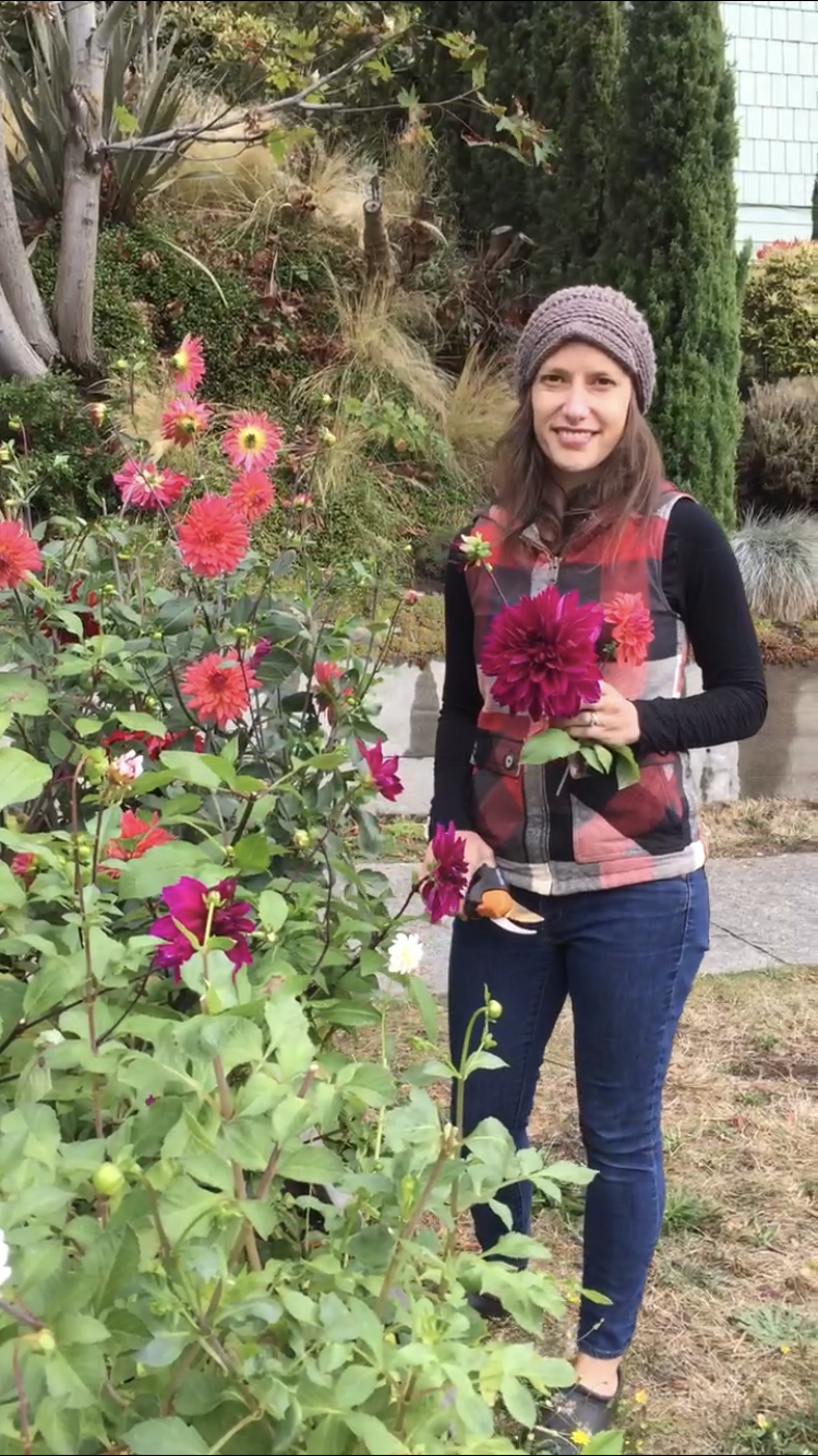 A woman standing outdoors in a garden, holding a large dark pink flower in one hand and a smaller flower in the other. She is dressed in a black long-sleeve shirt, a checkered vest, jeans, and a knitted gray beanie. There are various flowers and green plants around her, with a mixture of trees and shrubs in the background.