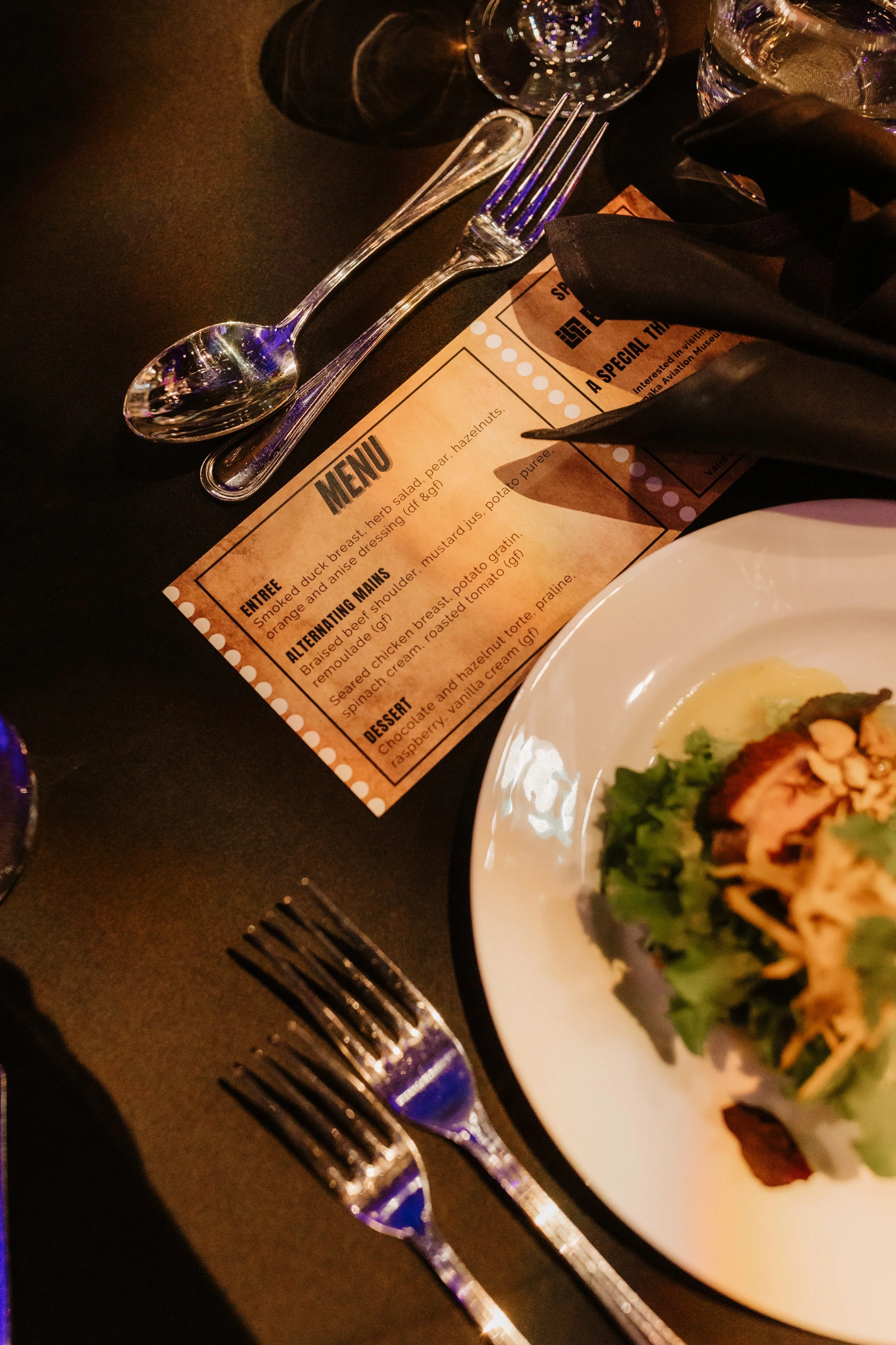 Partially visible salad on a white plate, a menu with a four-course meal, and two forks on a black table.