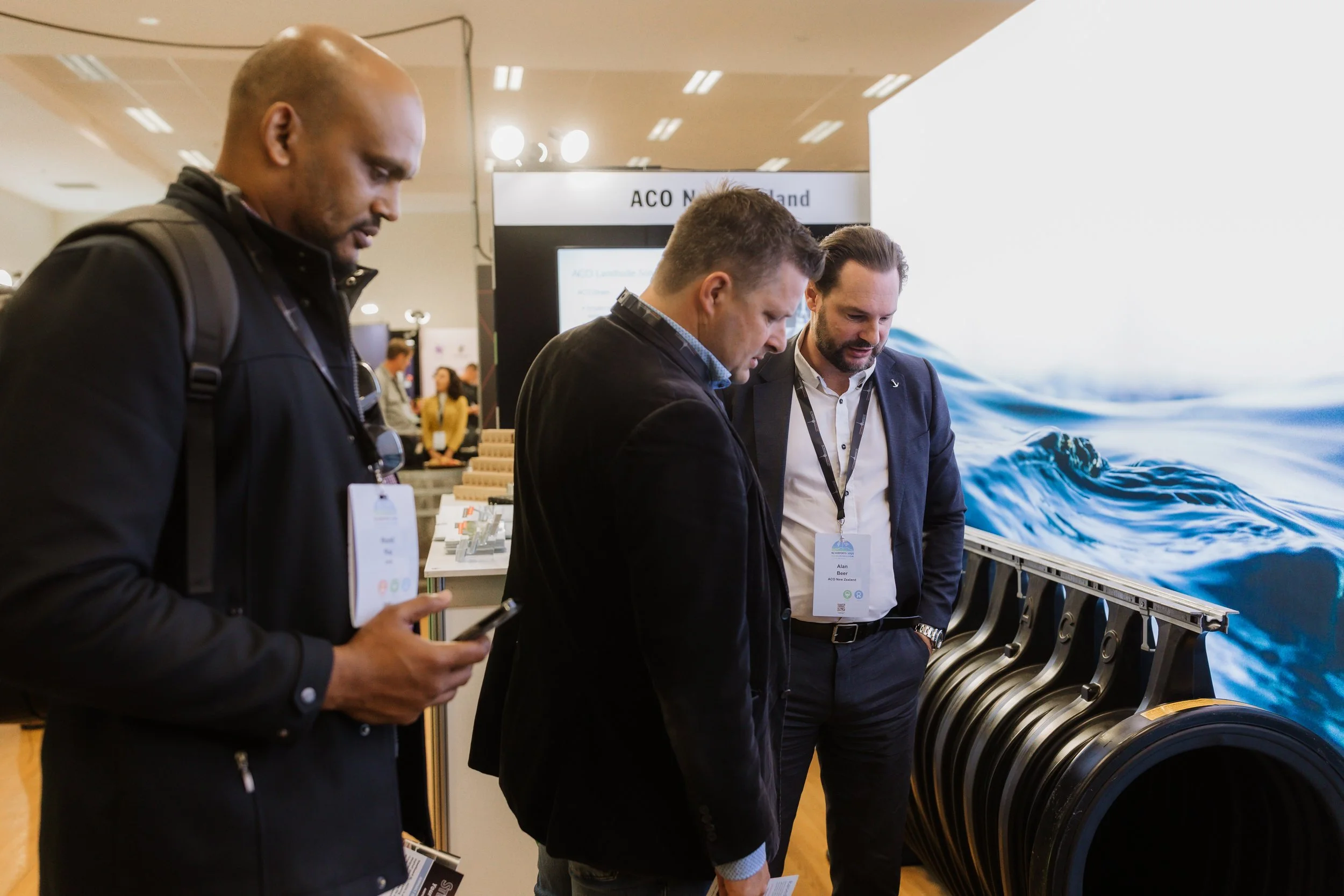 Three men examining a display at a trade show, with a large image of ocean waves in the background.