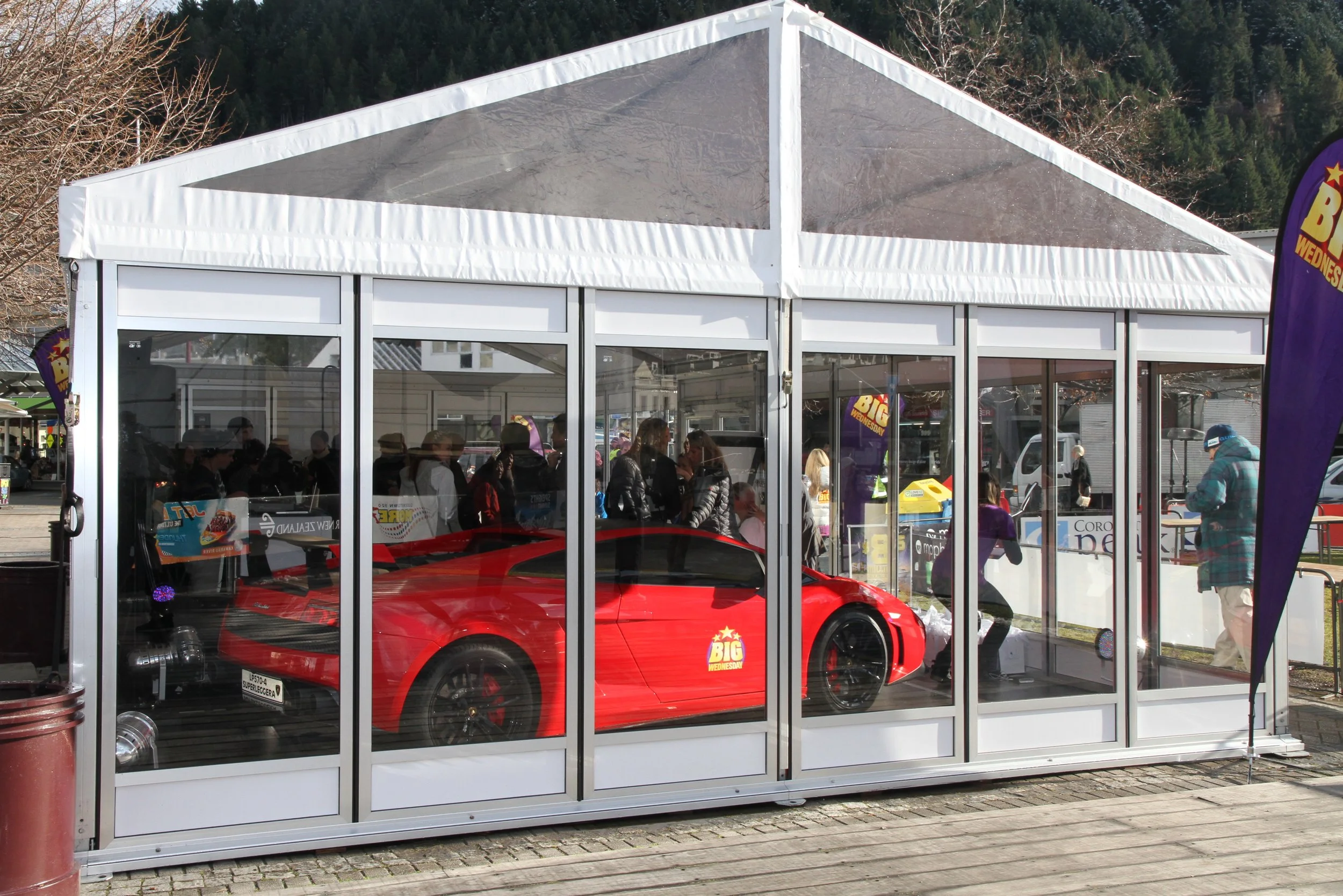 A red sports car inside a clear glass display enclosure at an outdoor event, with several people walking around and some purple flags visible.
